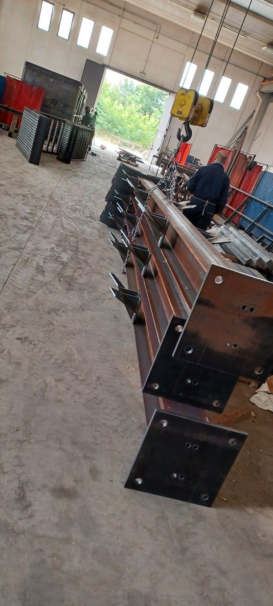 In un magazzino, una lunga trave metallica con piastre nere, sullo sfondo una persona in piedi vicino al gancio di una gru sospesa.