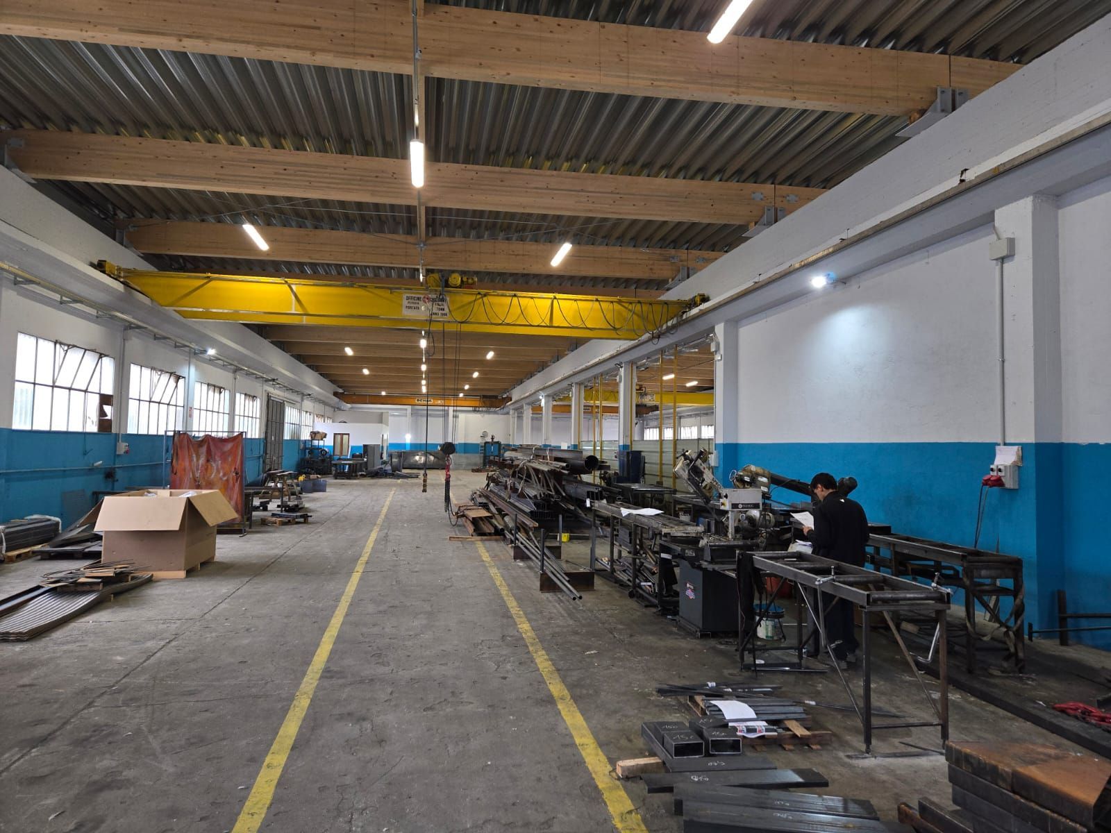 Un lungo pavimento di un magazzino industriale con una gru a ponte gialla, macchinari e una persona al lavoro in una postazione.