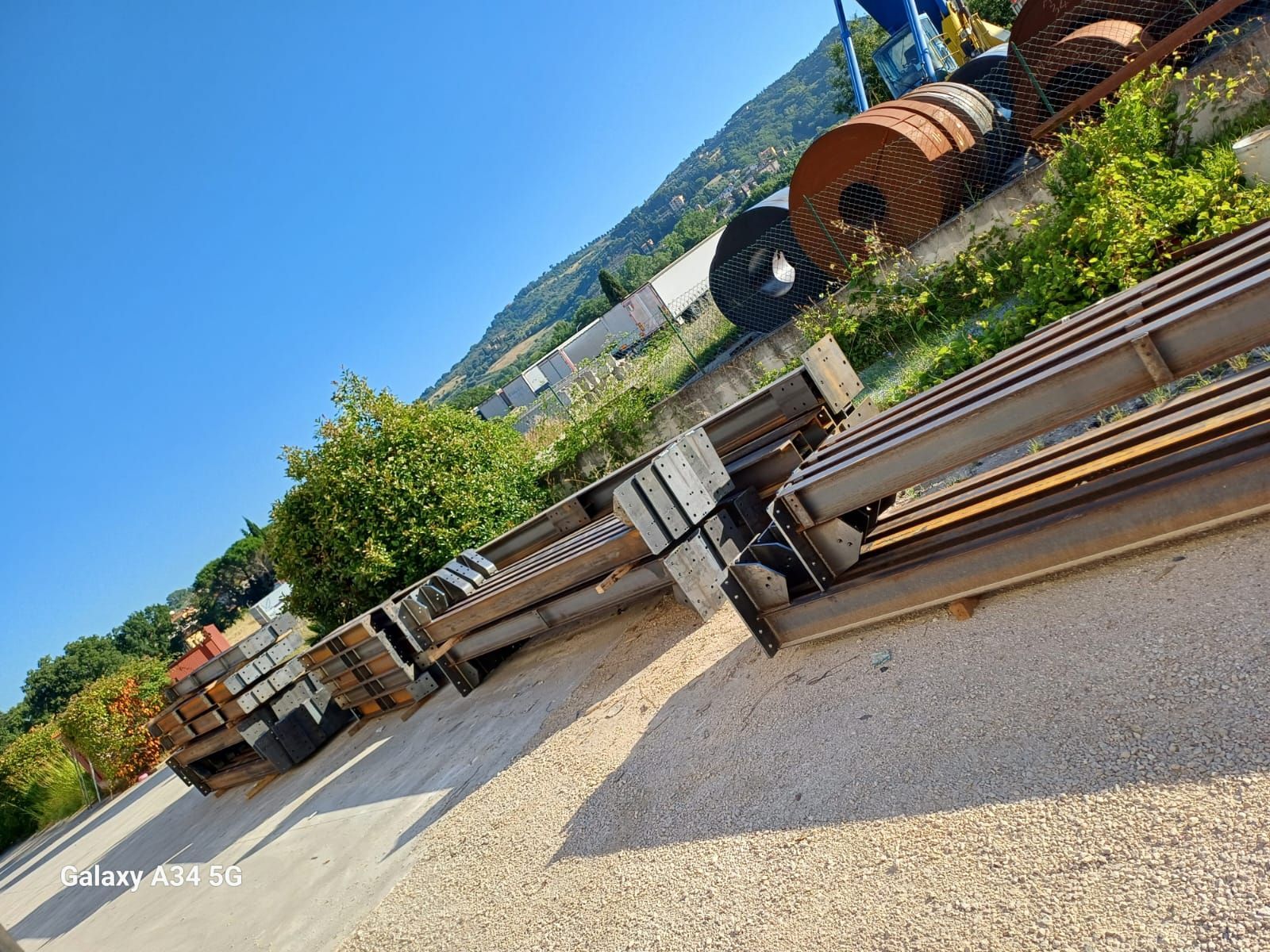 Travi e bobine d'acciaio impilate si trovano su un piazzale di ghiaia sotto un cielo azzurro brillante, con una rigogliosa collina verde in lontananza.