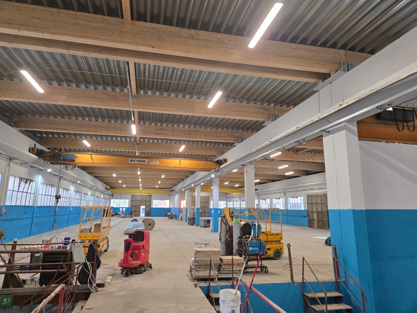 L'interno di un ampio e luminoso magazzino industriale è caratterizzato da travi in ​​legno, una gru a ponte gialla e attrezzi da costruzione.