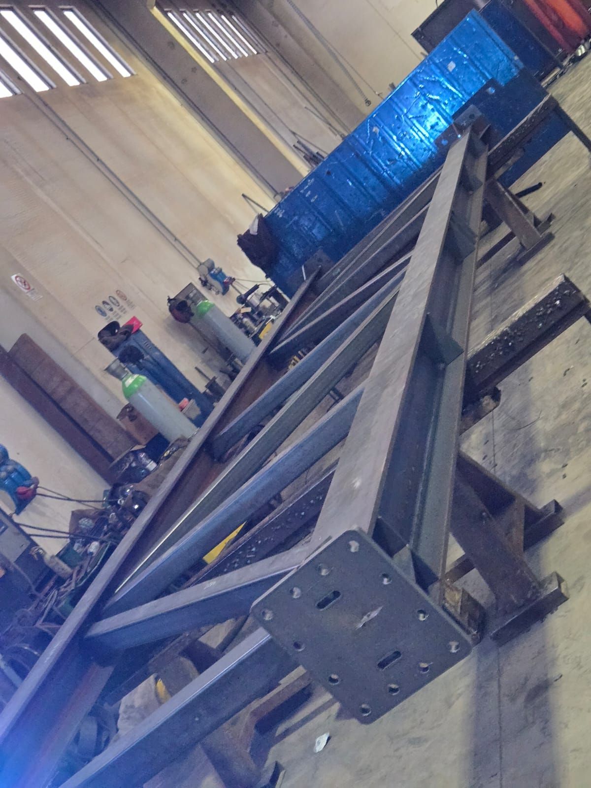 Una lunga trave reticolare in acciaio grigio con una piastra di montaggio quadrata all'estremità, appoggiata su dei supporti in un magazzino industriale.