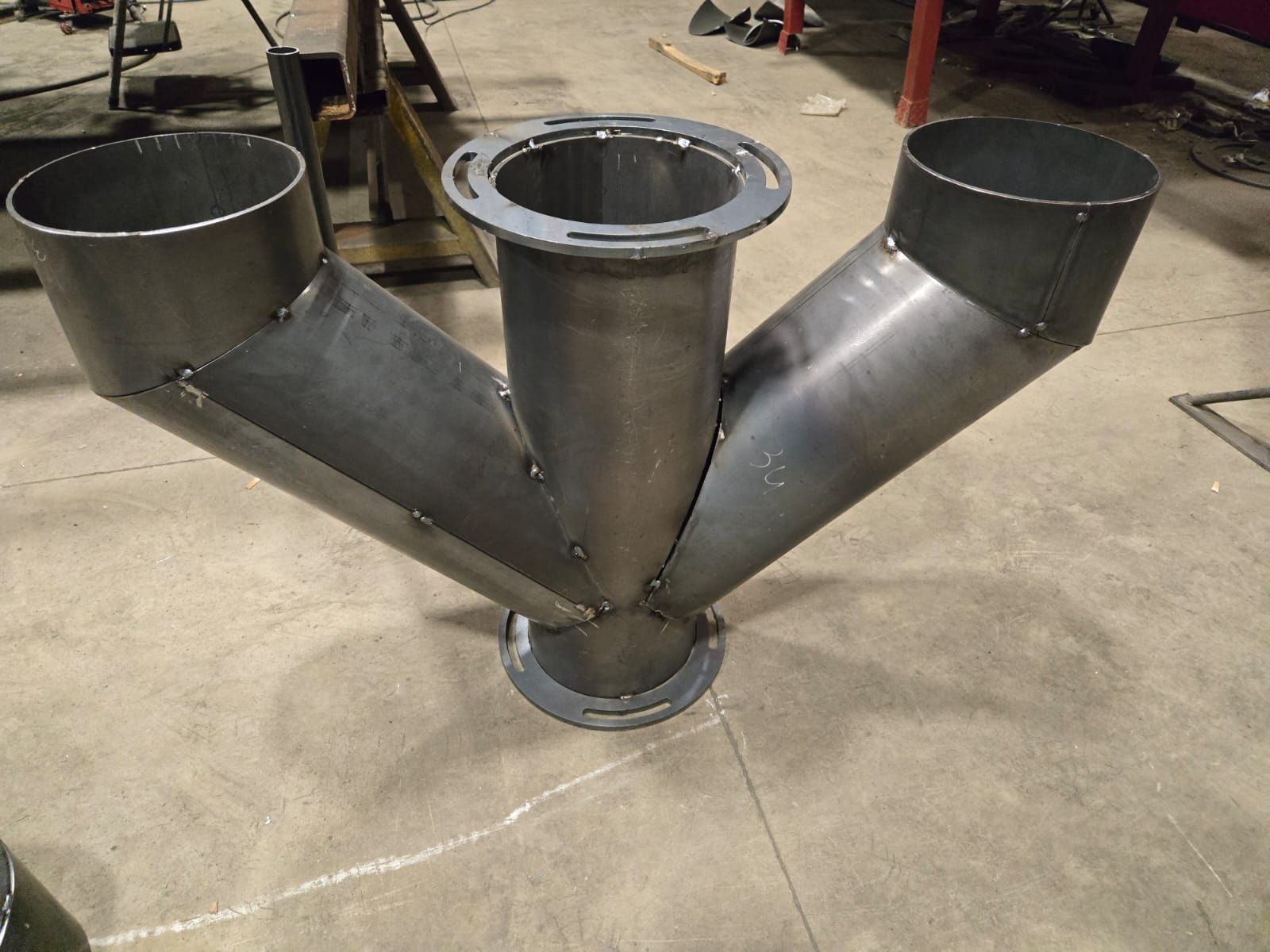 Un raccordo per tubi metallici a forma di Y con tre aperture circolari e flange saldate, posizionato su un pavimento di cemento di un'officina.