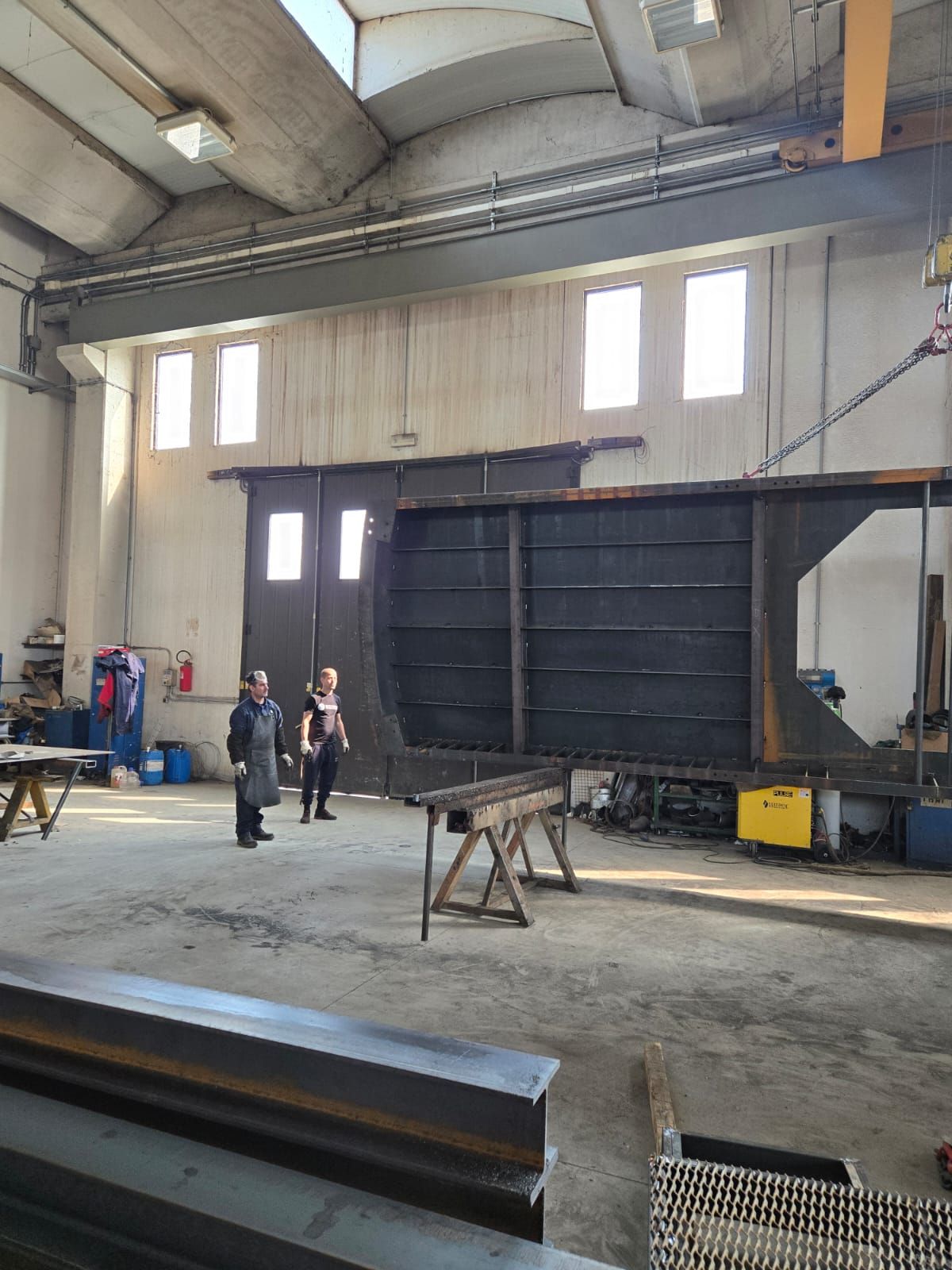 Due persone sono in piedi in una grande officina industriale, vicino a una struttura metallica sospesa e a un banco da lavoro.