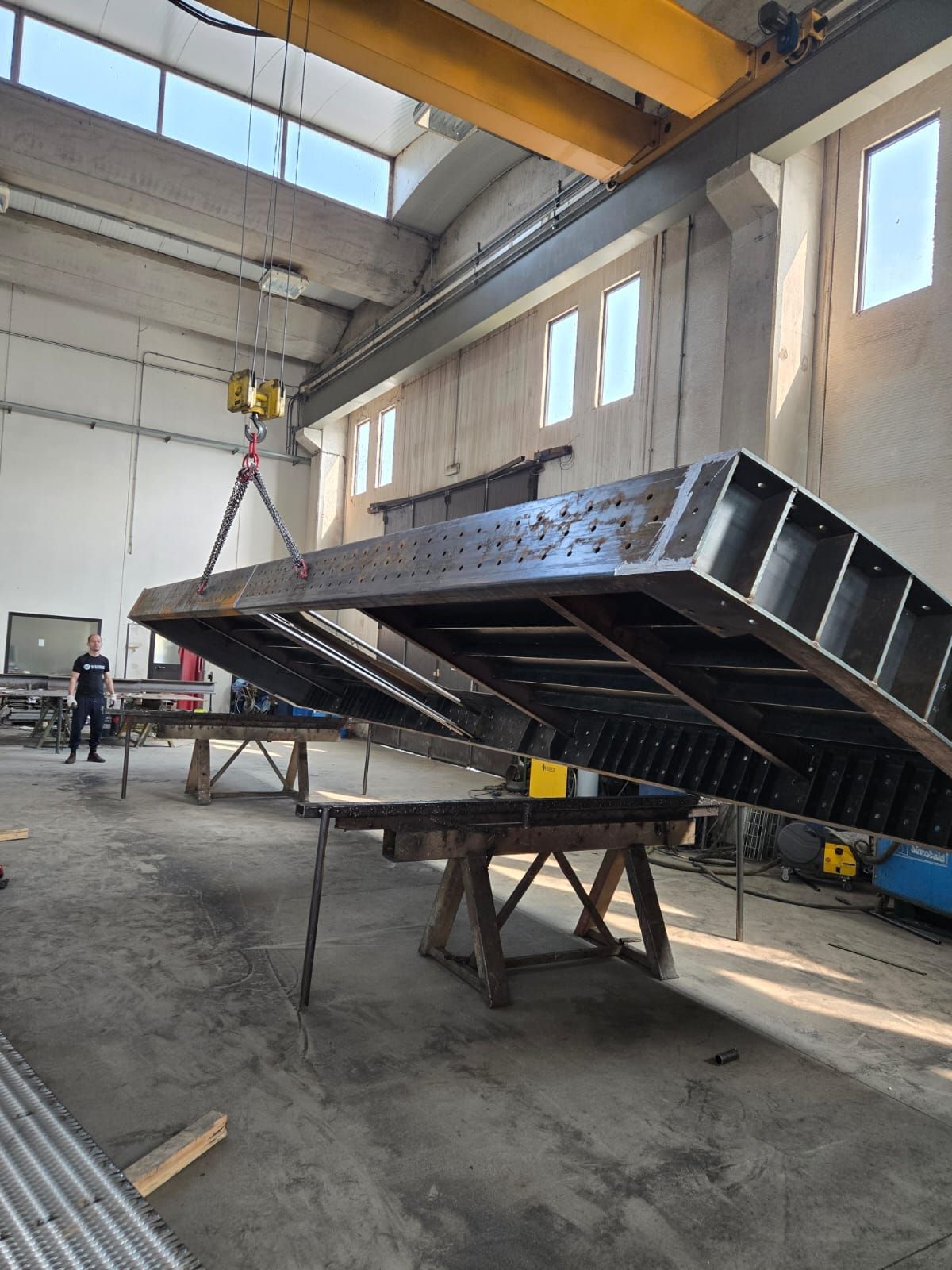 All'interno di un'officina, una gru solleva una grande struttura industriale in metallo scuro, sostenuta da cavalletti.