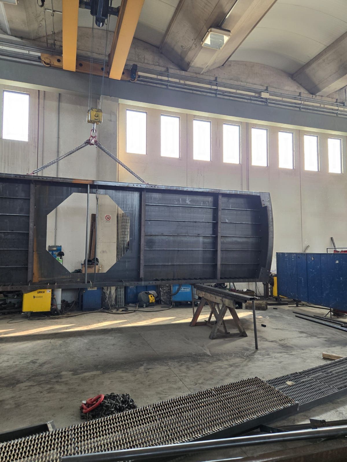 All'interno di un'officina, una grande struttura industriale in metallo scuro è sospesa tramite catene a una gru a ponte.