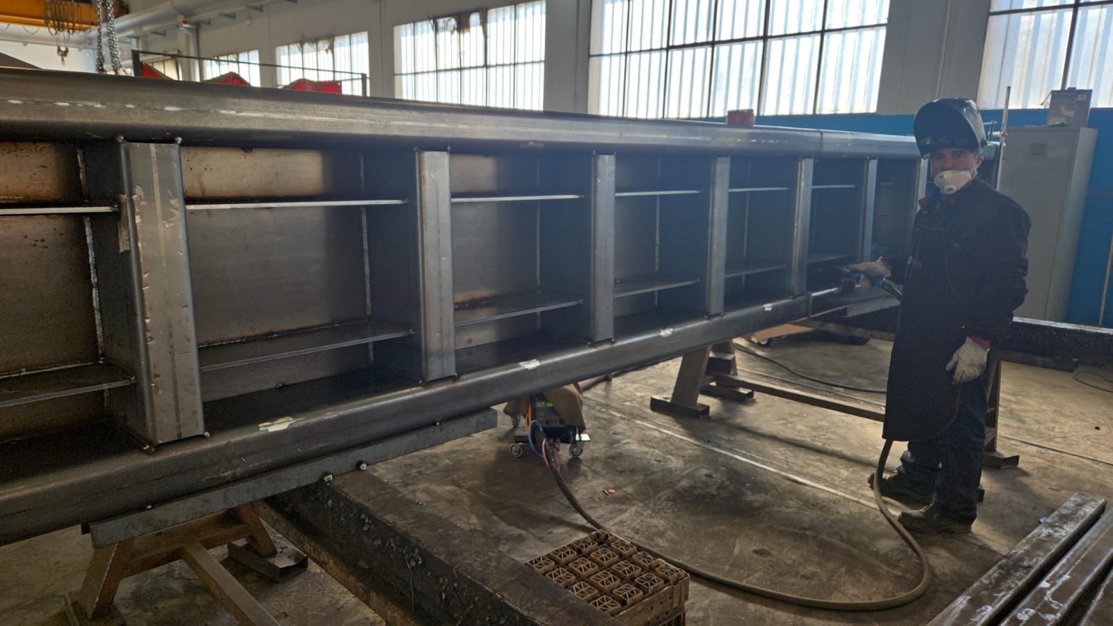 Un saldatore con indumenti protettivi è in piedi accanto a una grande trave d'acciaio industriale in un'officina.