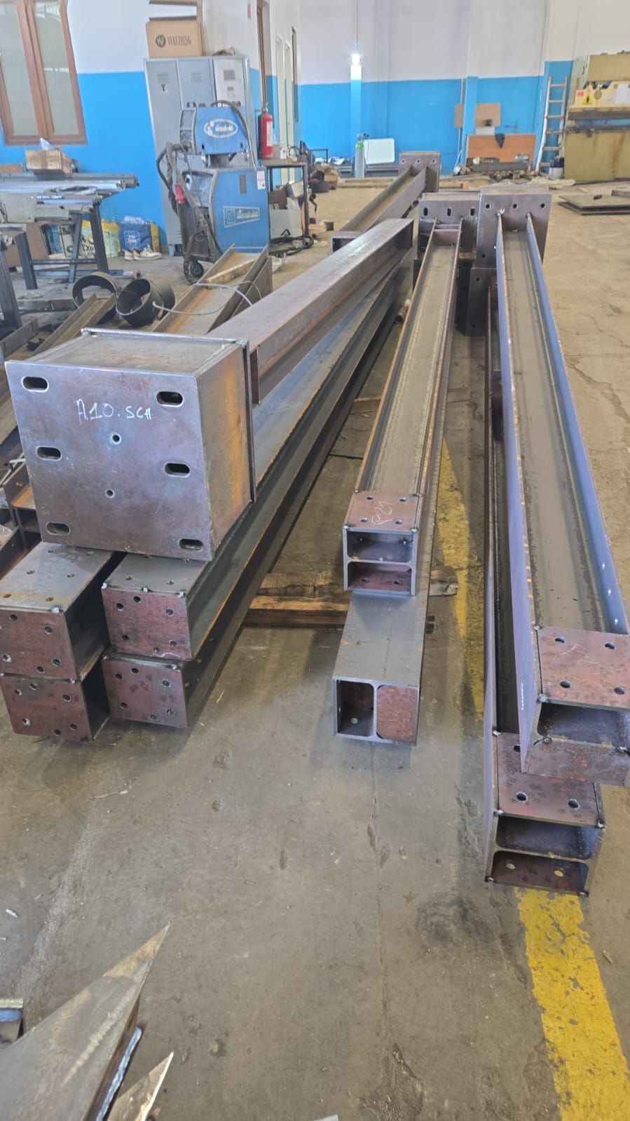 Diverse lunghe travi a H in metallo grigio, con piastre terminali saldate, poggiano sul pavimento di cemento di un'officina di fabbricazione.