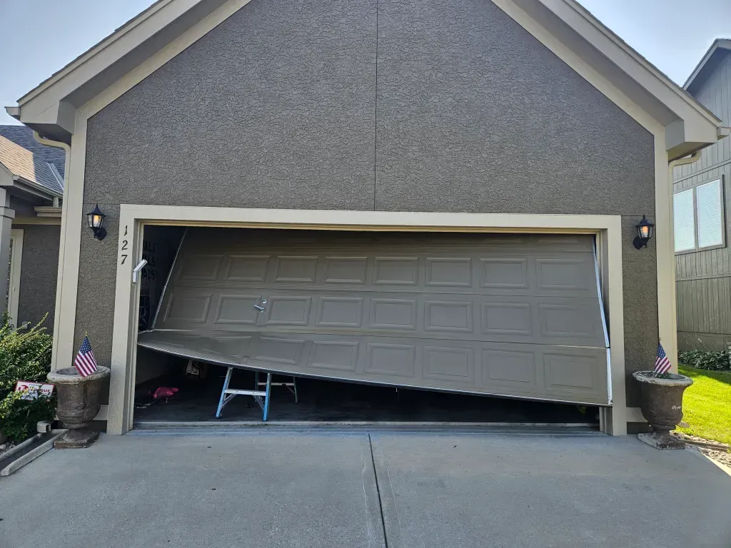 Damaged garage door, partially open. The door is bent and askew. A small stepladder is visible inside the garage.