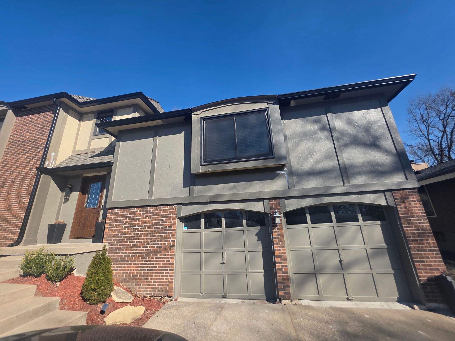 Two-story house with gray siding, brick accents, and a two-car garage under a blue sky.