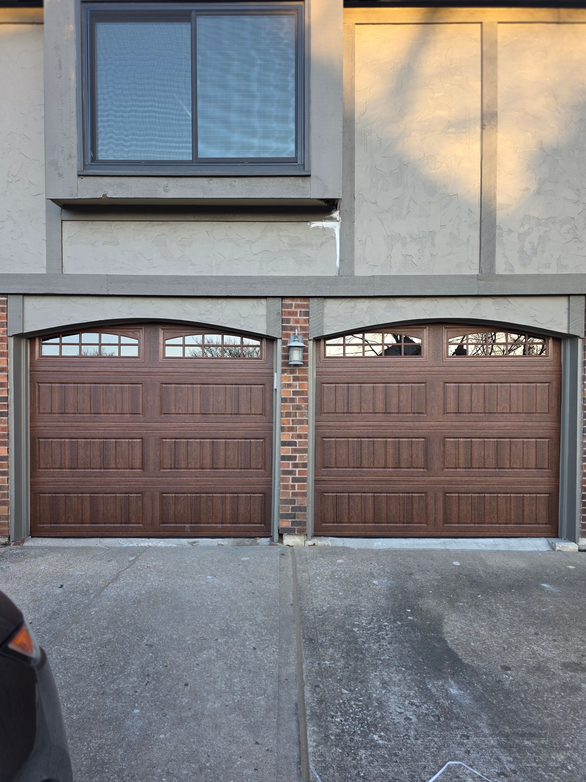 Two brown garage doors with arched top windows, brick accents, and a window above.