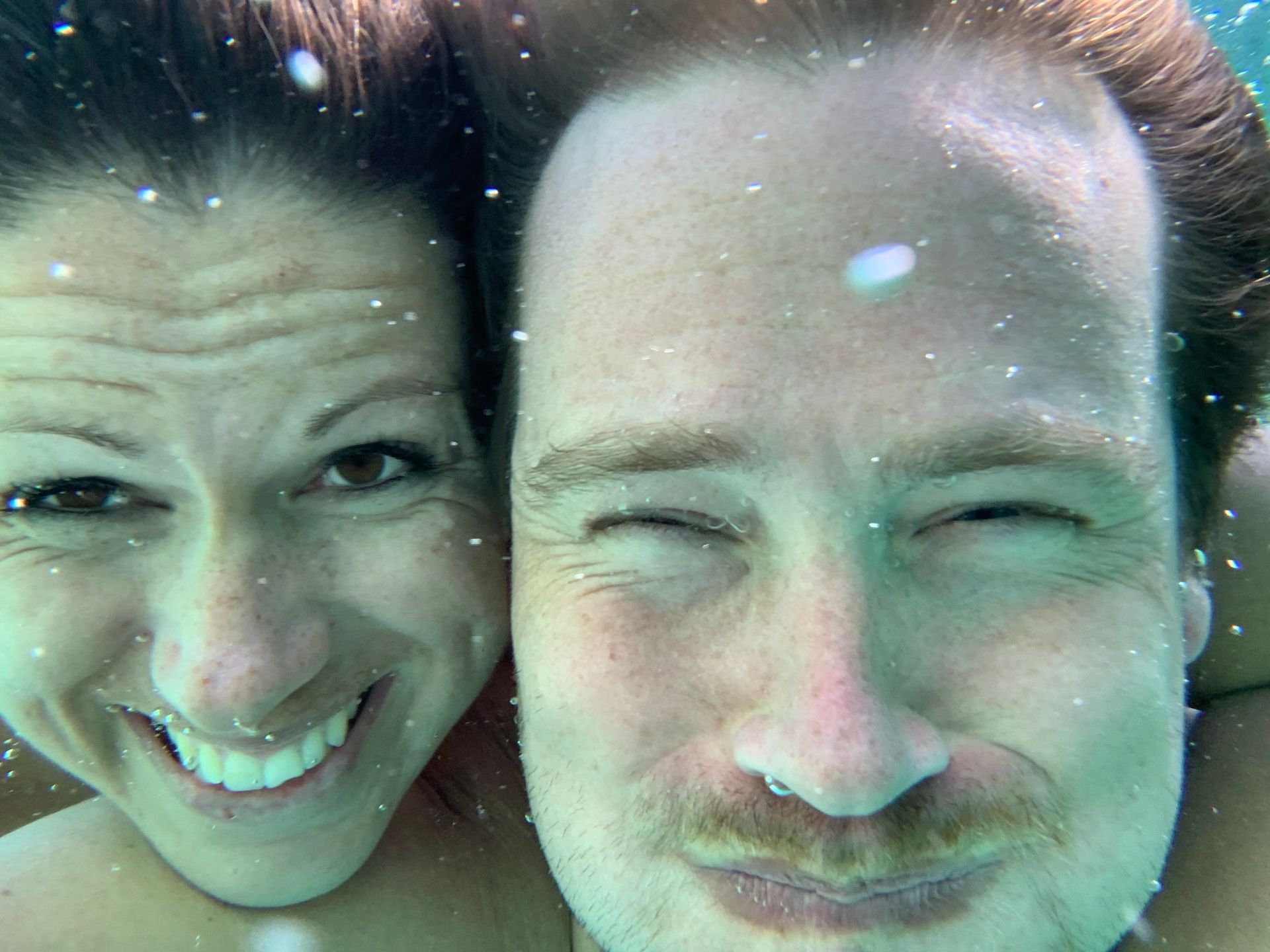 Couple underwater, smiling, one winking, with sunlight, turquoise water.