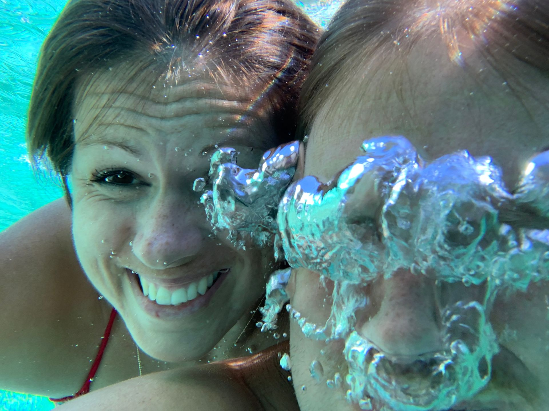 Two women underwater, one smiling, the other blowing bubbles in a pool.