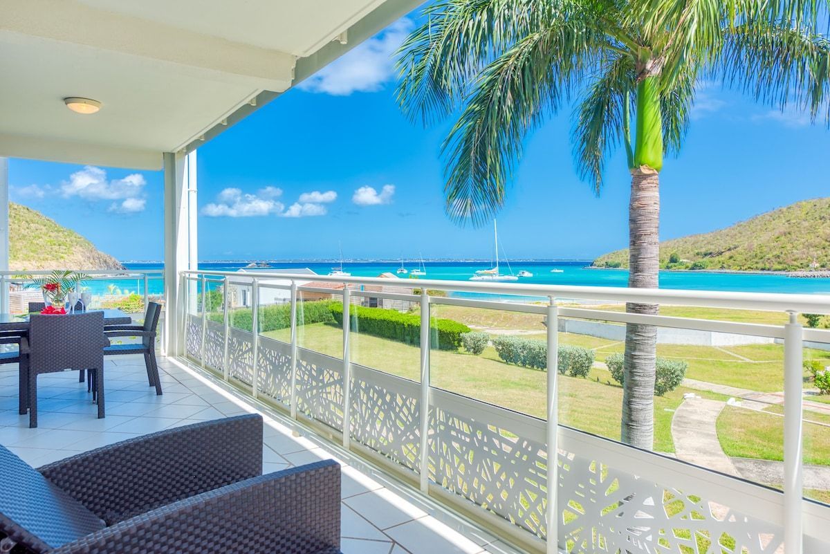 Balcony overlooking turquoise water, palm tree, boats, and blue sky.