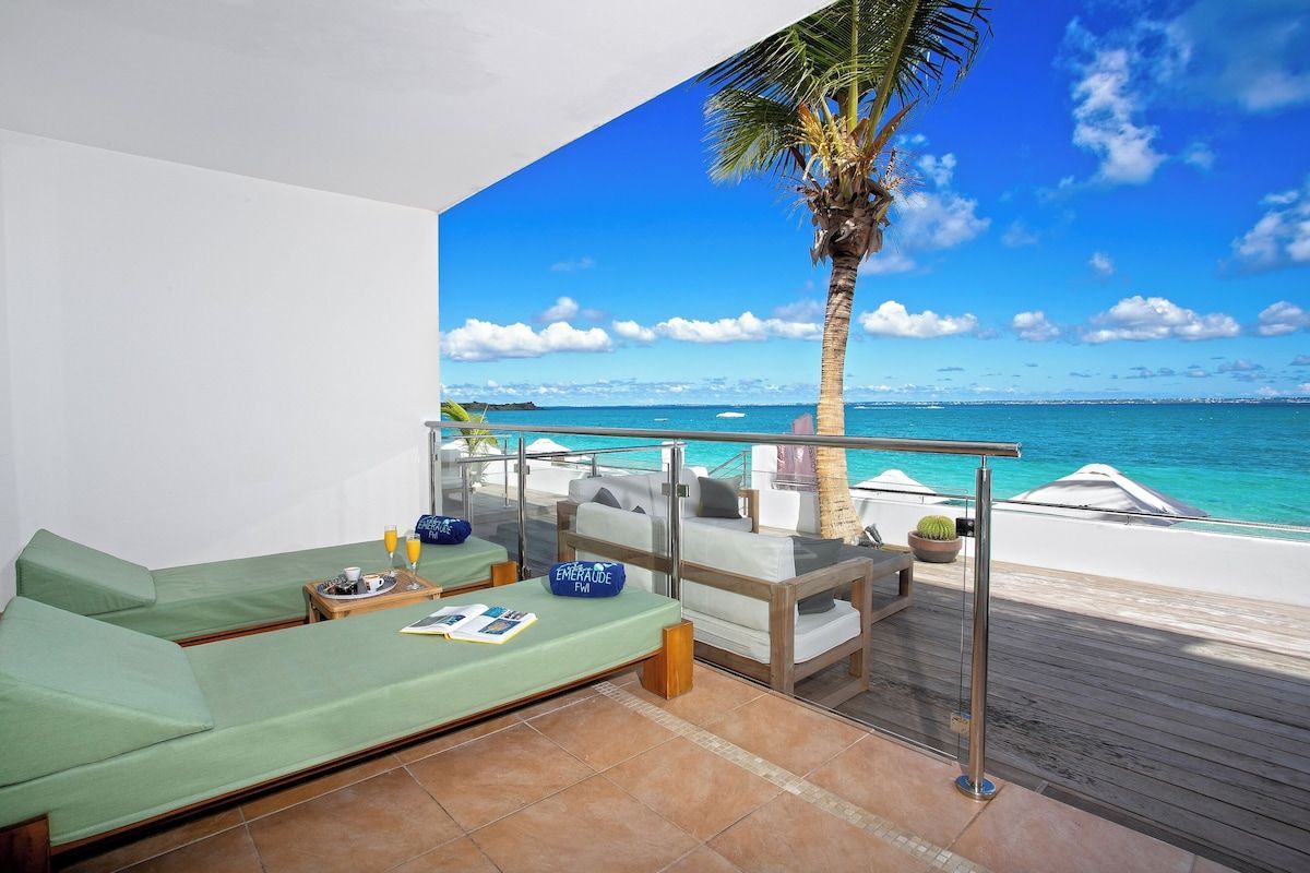 Balcony overlooking a beach with lounge chairs, ocean, and palm tree under a bright blue sky.