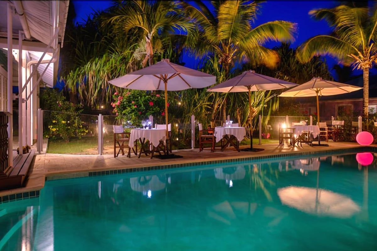 Poolside restaurant with white tables, umbrellas, and palm trees illuminated at night.
