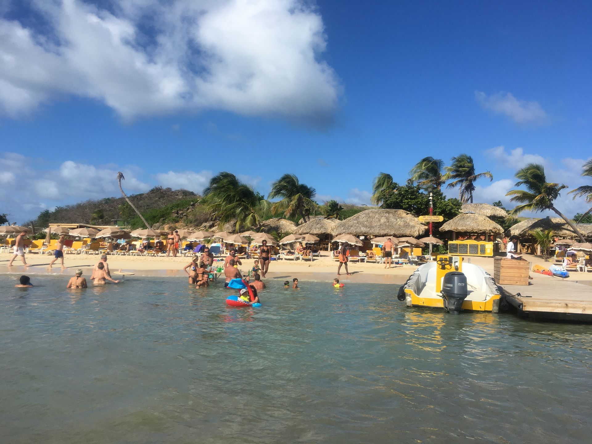 Beach scene: people swimming and relaxing on the shore with thatched-roof huts and palm trees under a blue sky.