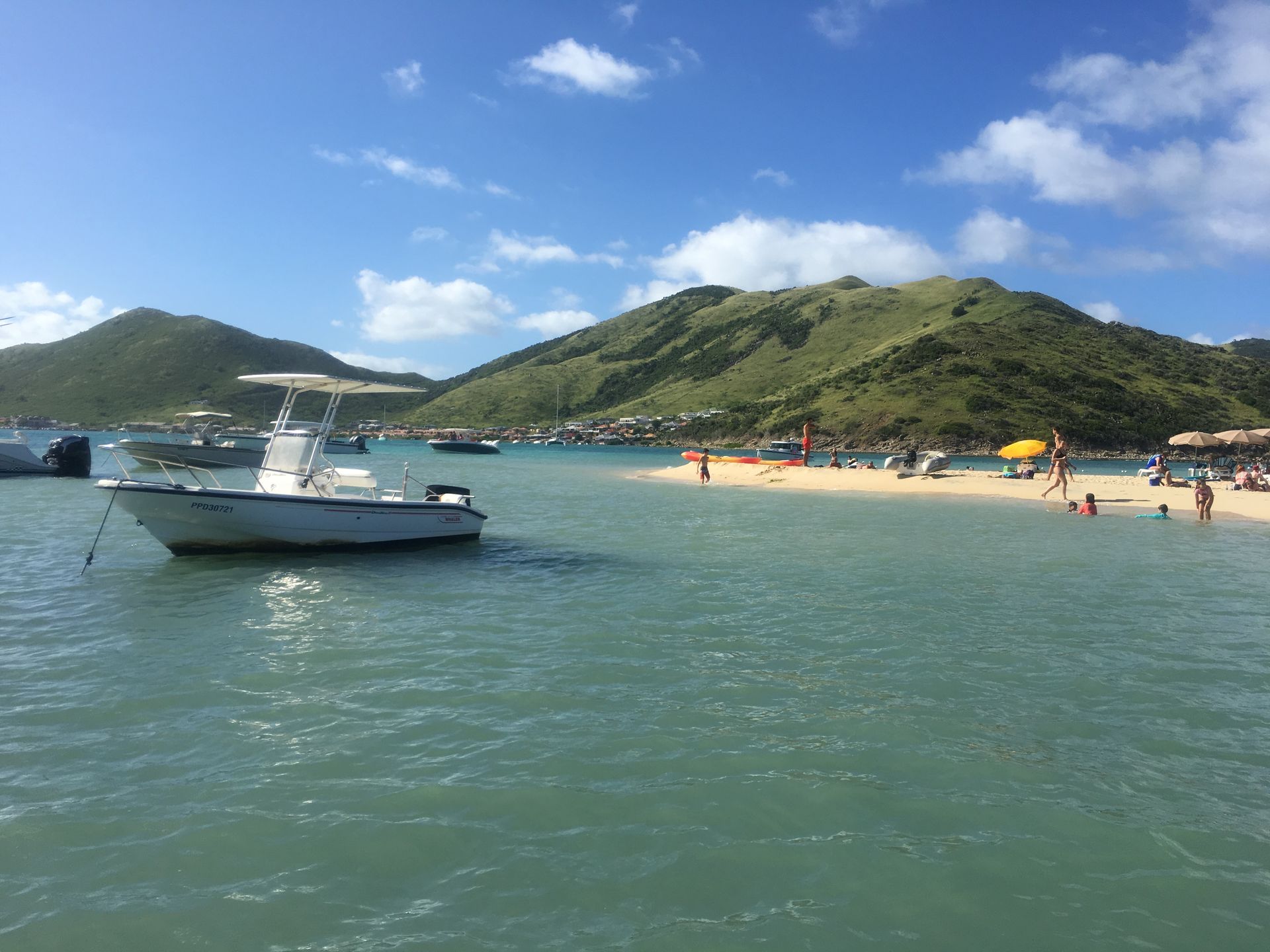 White boat in turquoise water near a sandy beach with people. Green hills and blue sky in the background.