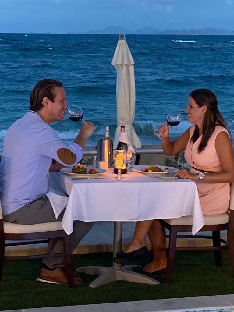 Couple toasting wine at a seaside dinner table at dusk; woman in peach dress, man in light blue shirt.
