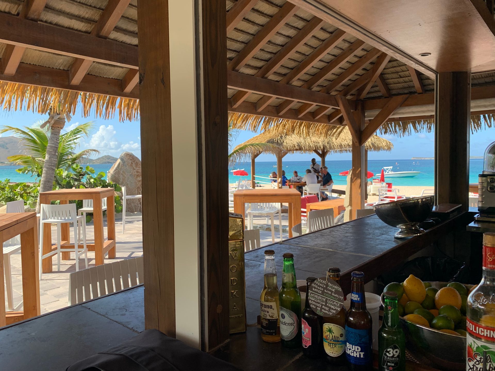 Beachside bar with a view of the ocean, people, and sandy beach. Bottles and limes on the bar.