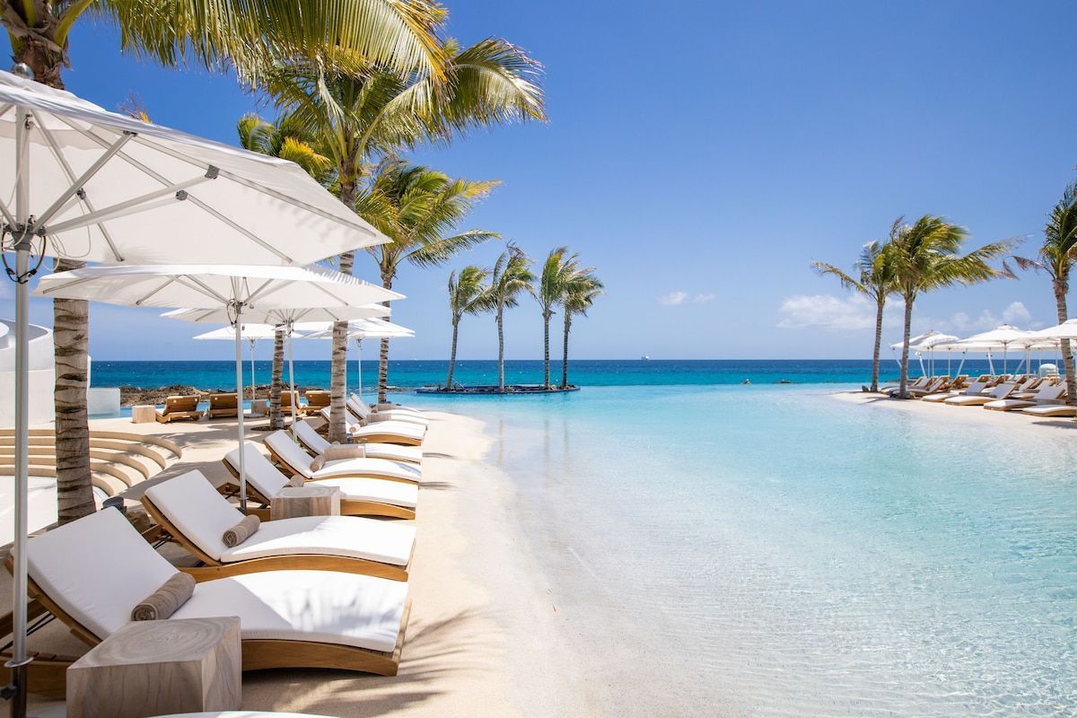 Luxury resort pool overlooking a turquoise ocean. White lounge chairs, umbrellas, and palm trees under a blue sky.