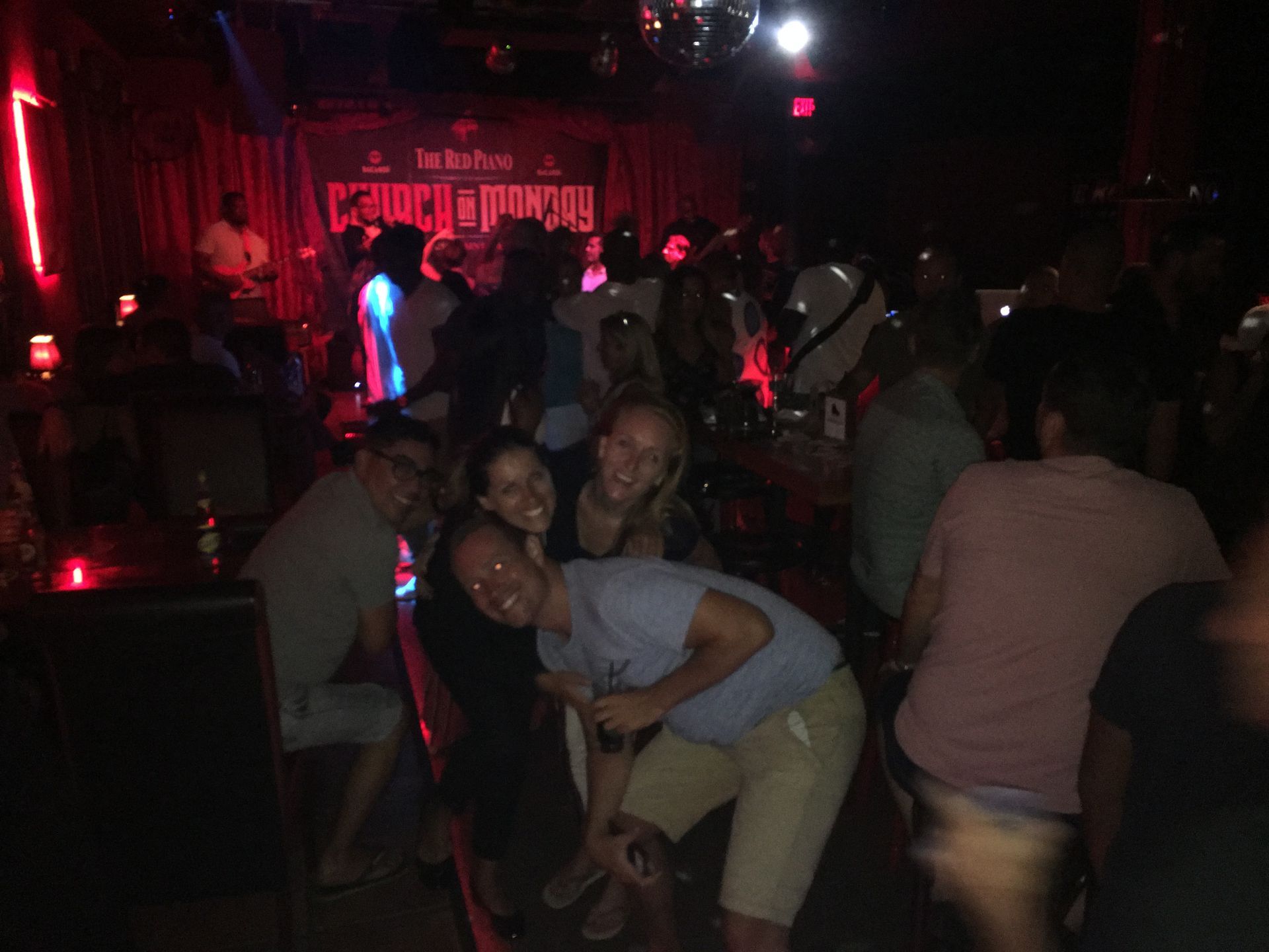 People gathered at a dimly lit bar, smiling. Red lighting, a disco ball, and a band visible in the background.