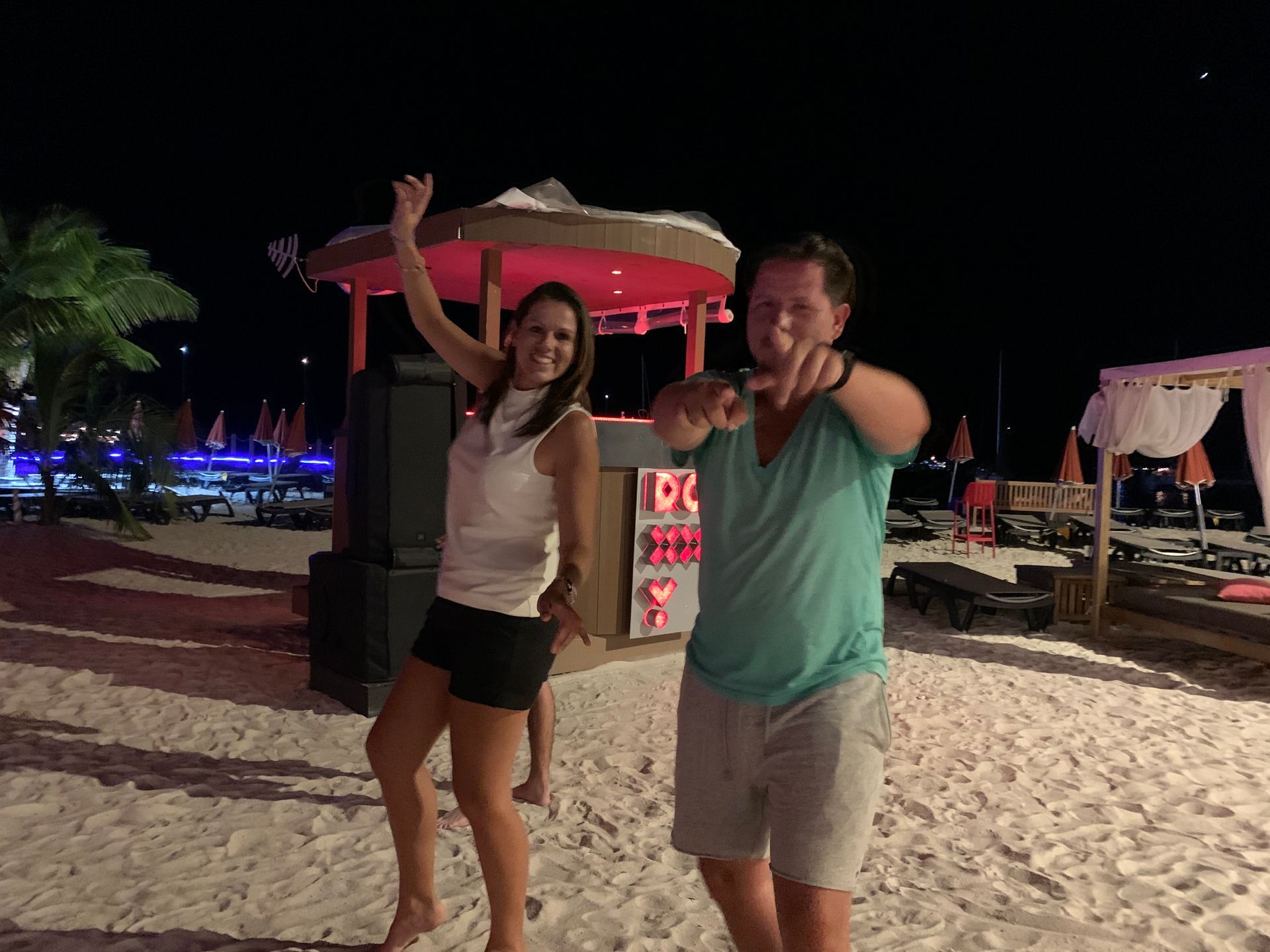 Woman and man dancing on a sandy beach at night; woman raises her hand; man points forward. Pink-lit bar in the background.
