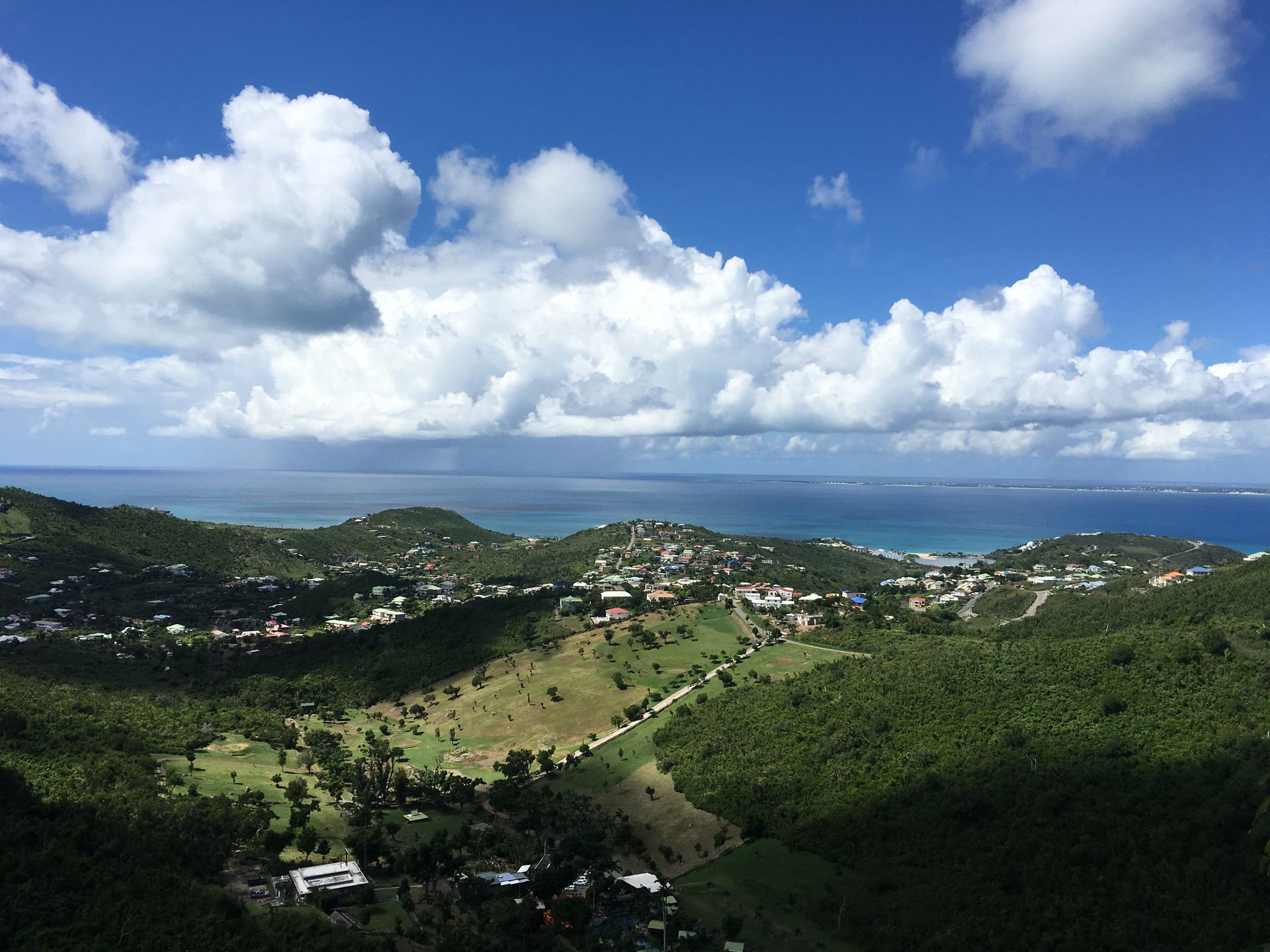 Green hills overlooking a coastal town and blue ocean under a partly cloudy sky.