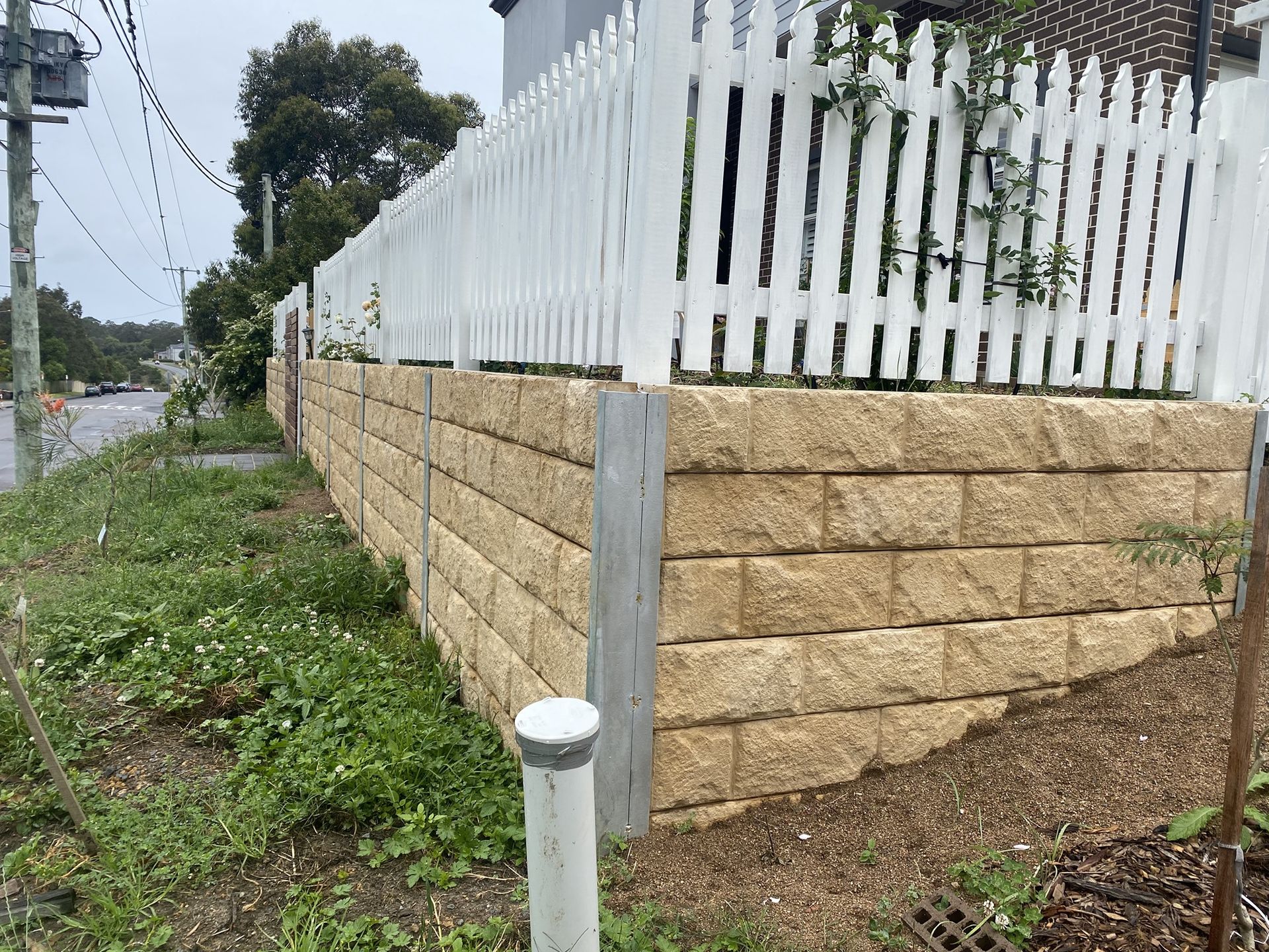 A Retaining Wall Made of Tan Blocks Supports a White Picket Fence — Concrete Sleepers NSW in Newcastle, NSW
