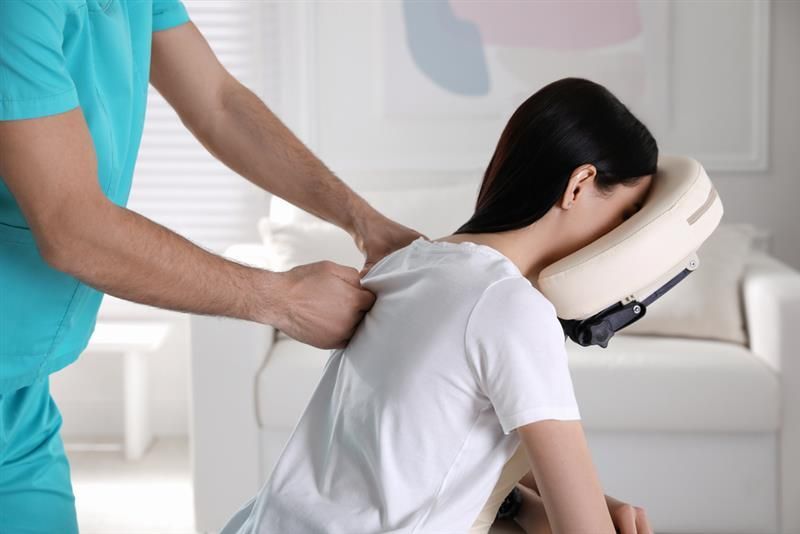 Person receiving a massage on a massage chair, indoors. Therapist in teal scrubs, white shirt, beige chair.
