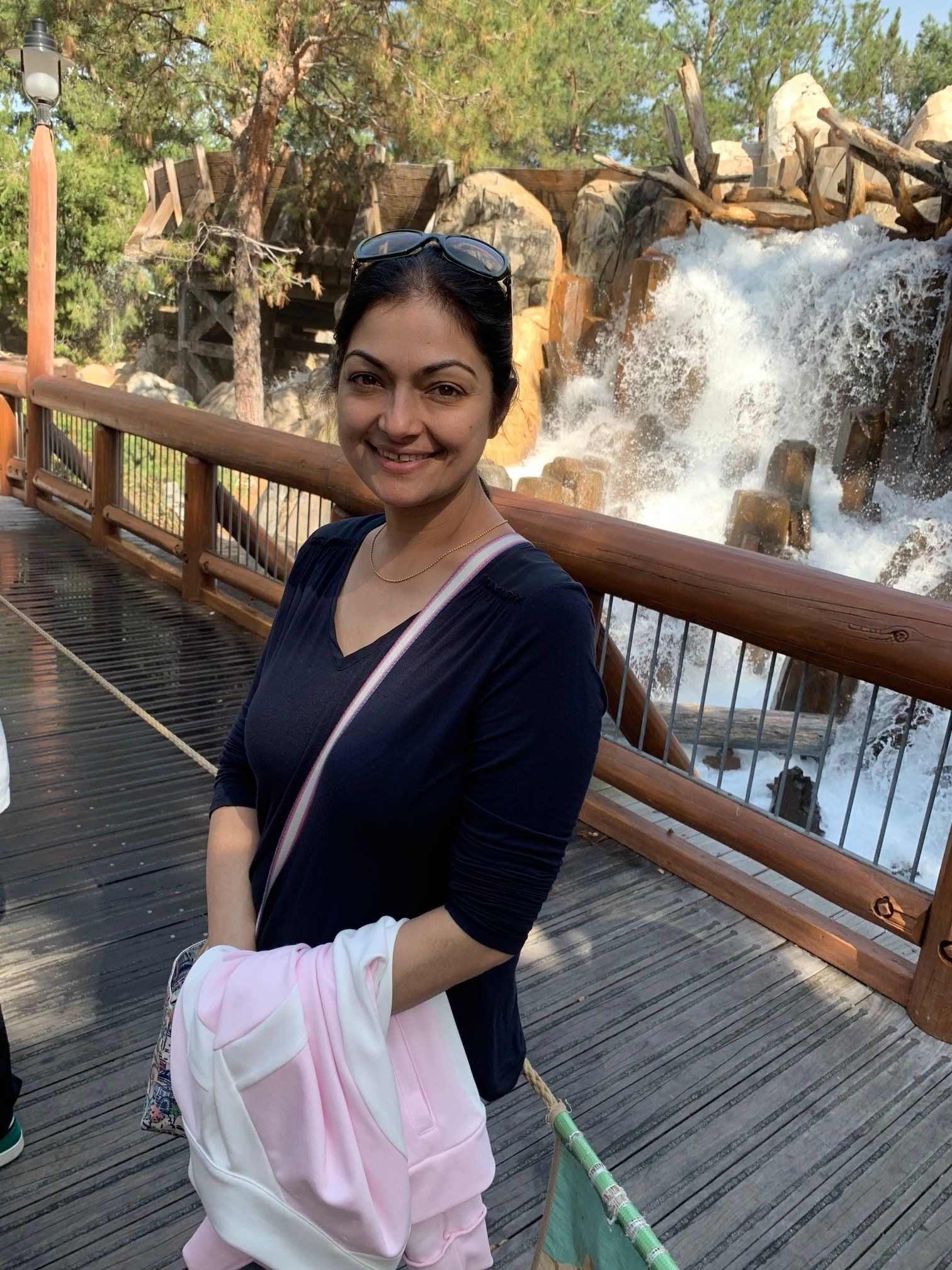 Woman smiling on a bridge with a waterfall backdrop. She is wearing a navy top, and holding a pink jacket.