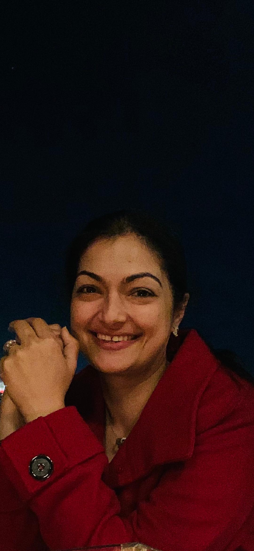 Smiling woman wearing a red coat, hands clasped in front of her. Dark blue background.
