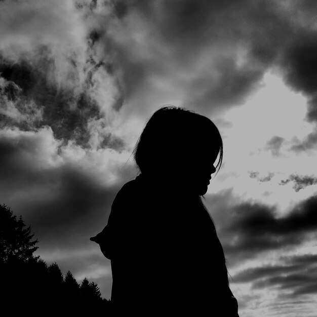 A black and white photo of a person standing in front of a cloudy sky.