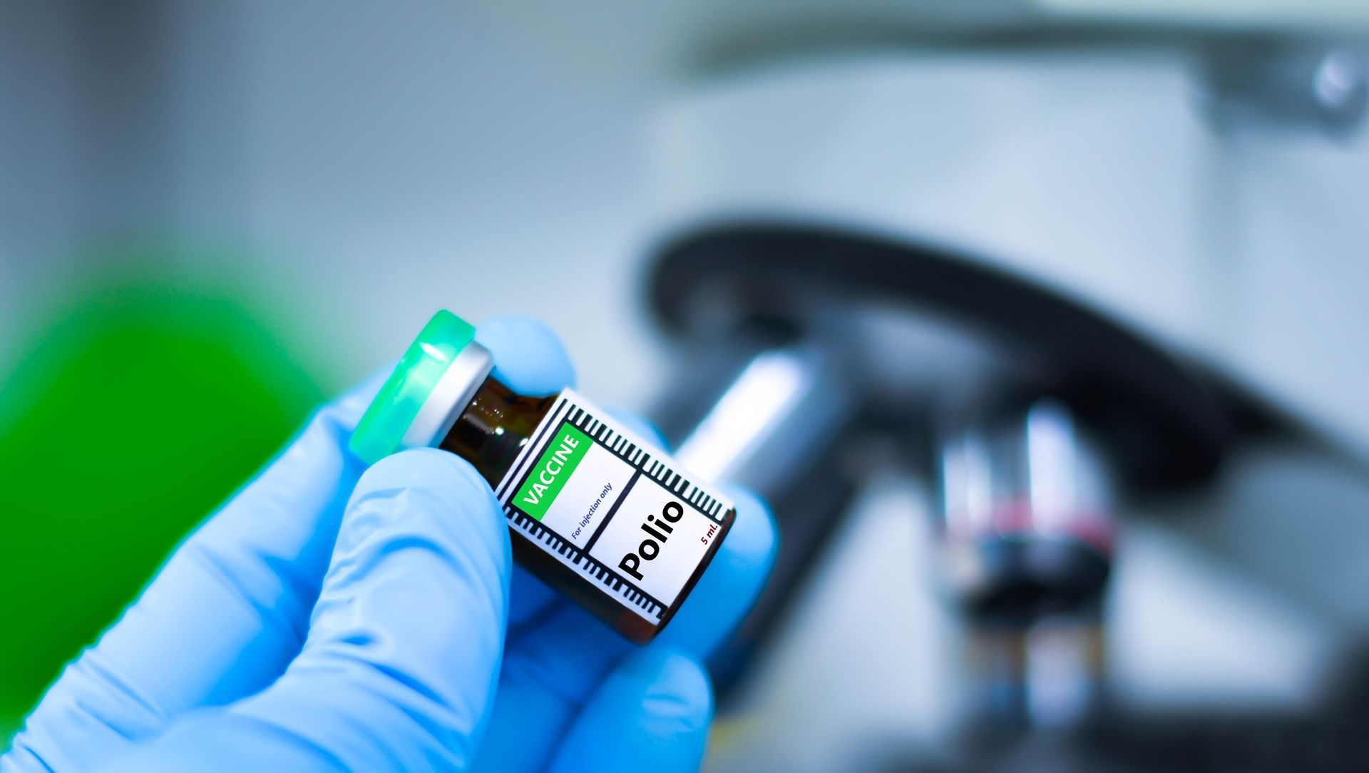 A person wearing blue gloves is holding a bottle of vaccine in front of a microscope.