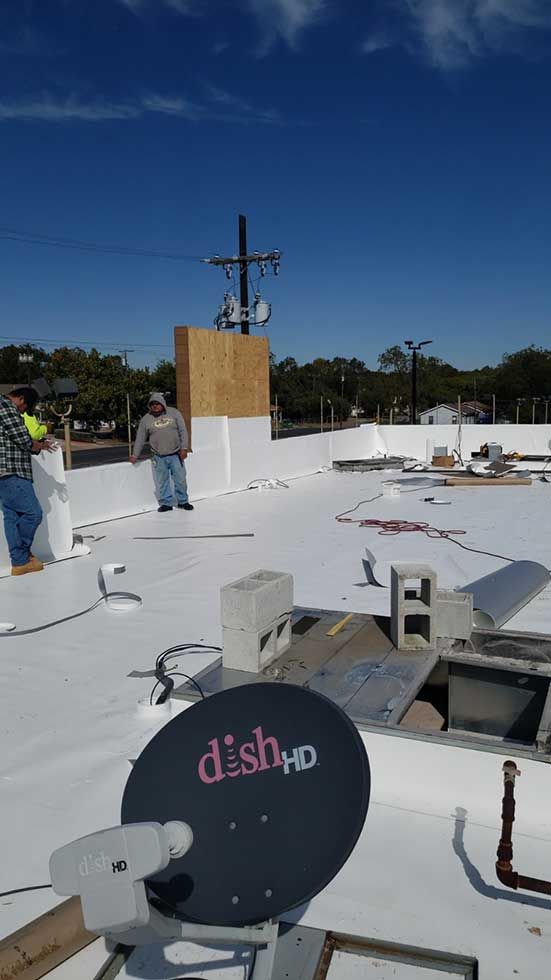 A man is standing on top of a white roof next to a dish tv satellite dish.