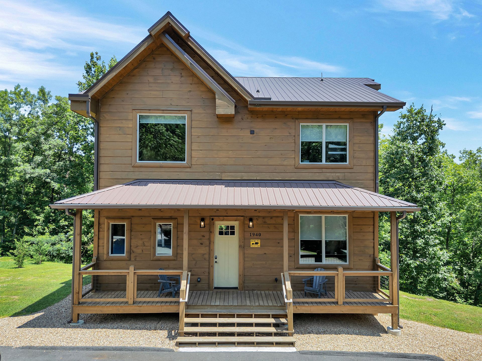 Wooden two-story cabin with porch and metal roof, set in a green, sunny environment.