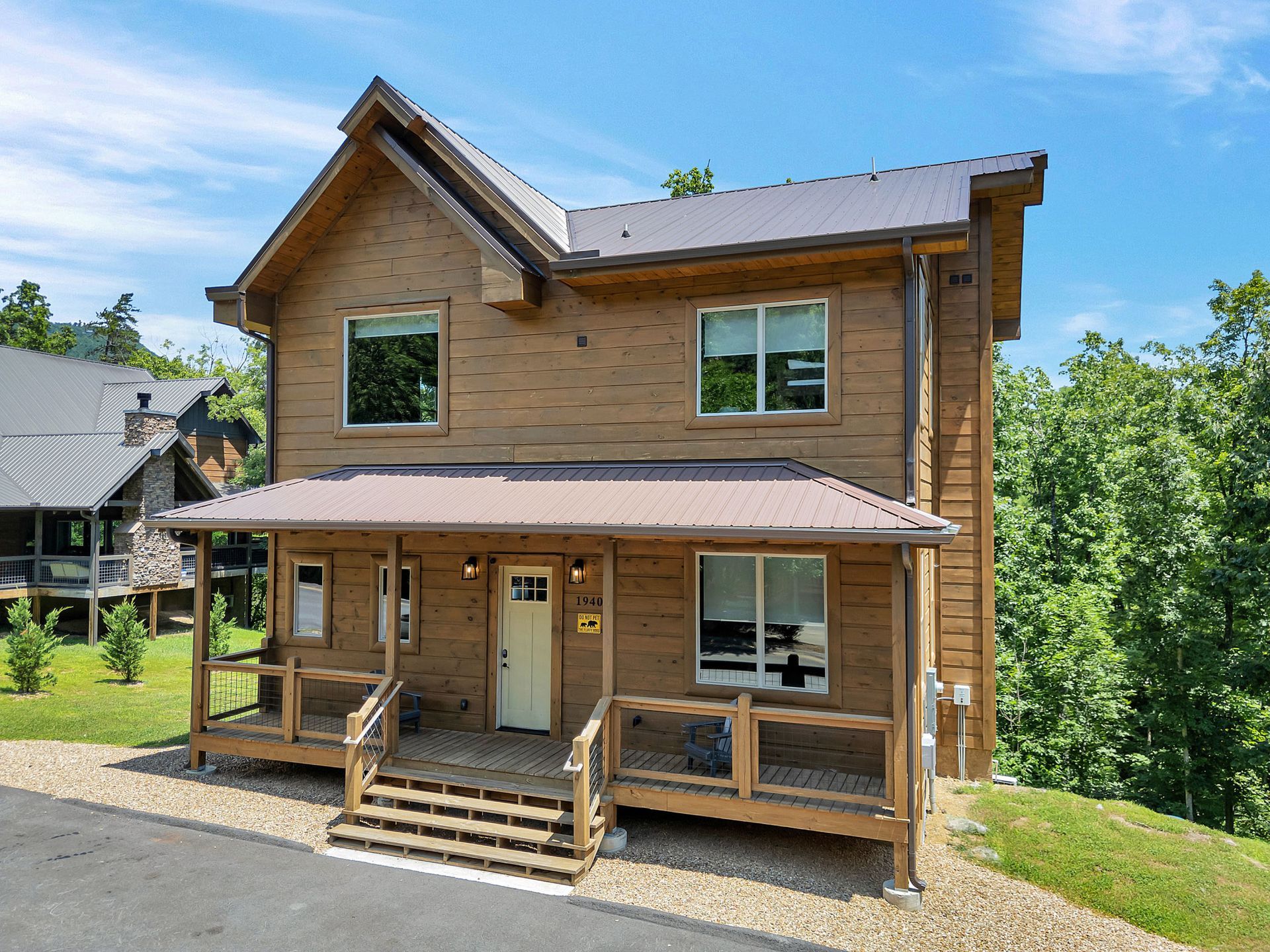 Two-story brown cabin with porch, steps, and metal roof, set among trees on a slight hill.