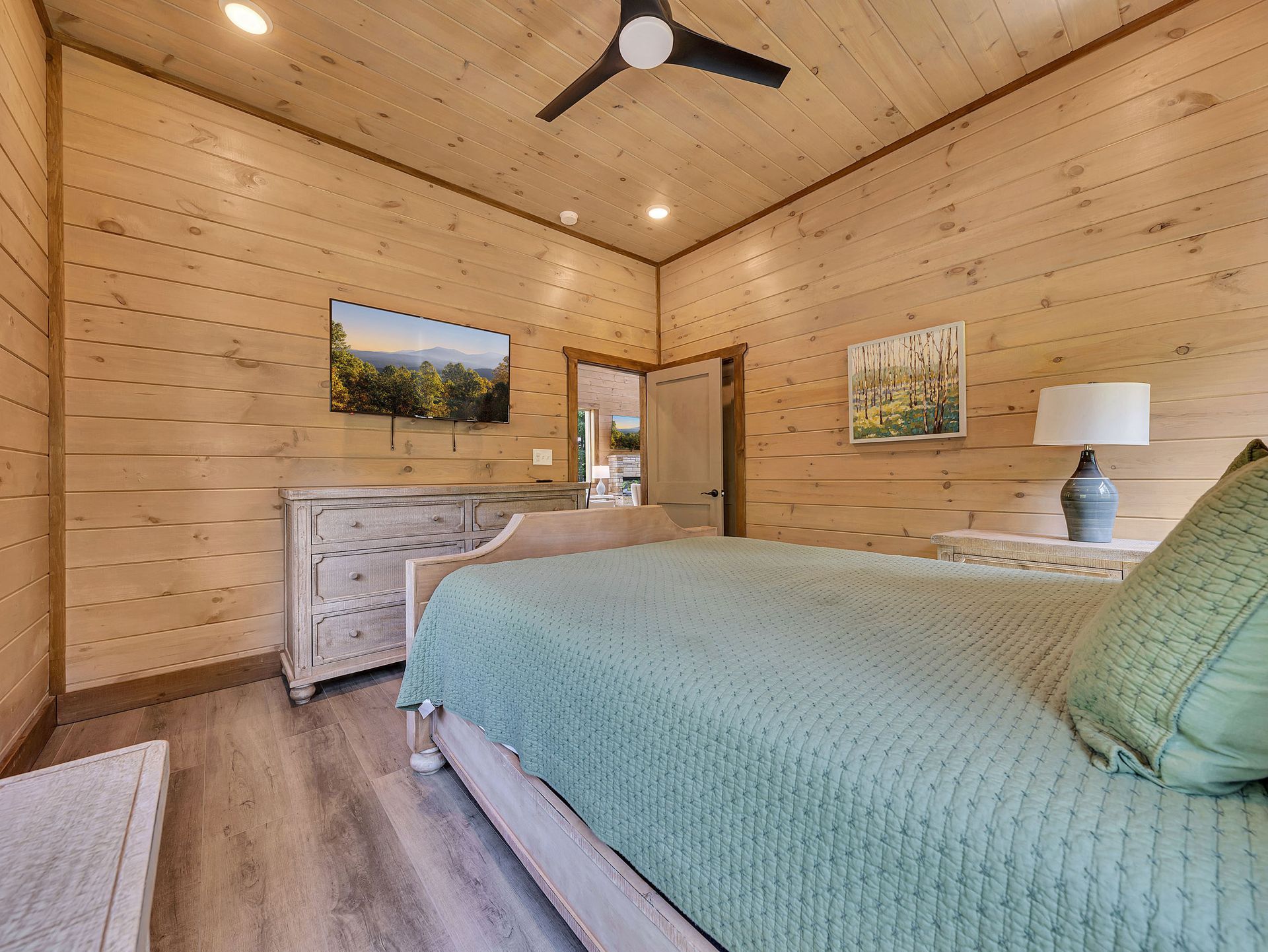 Bedroom with wood-paneled walls, bed with green comforter, TV, dresser, and side tables with lamps.