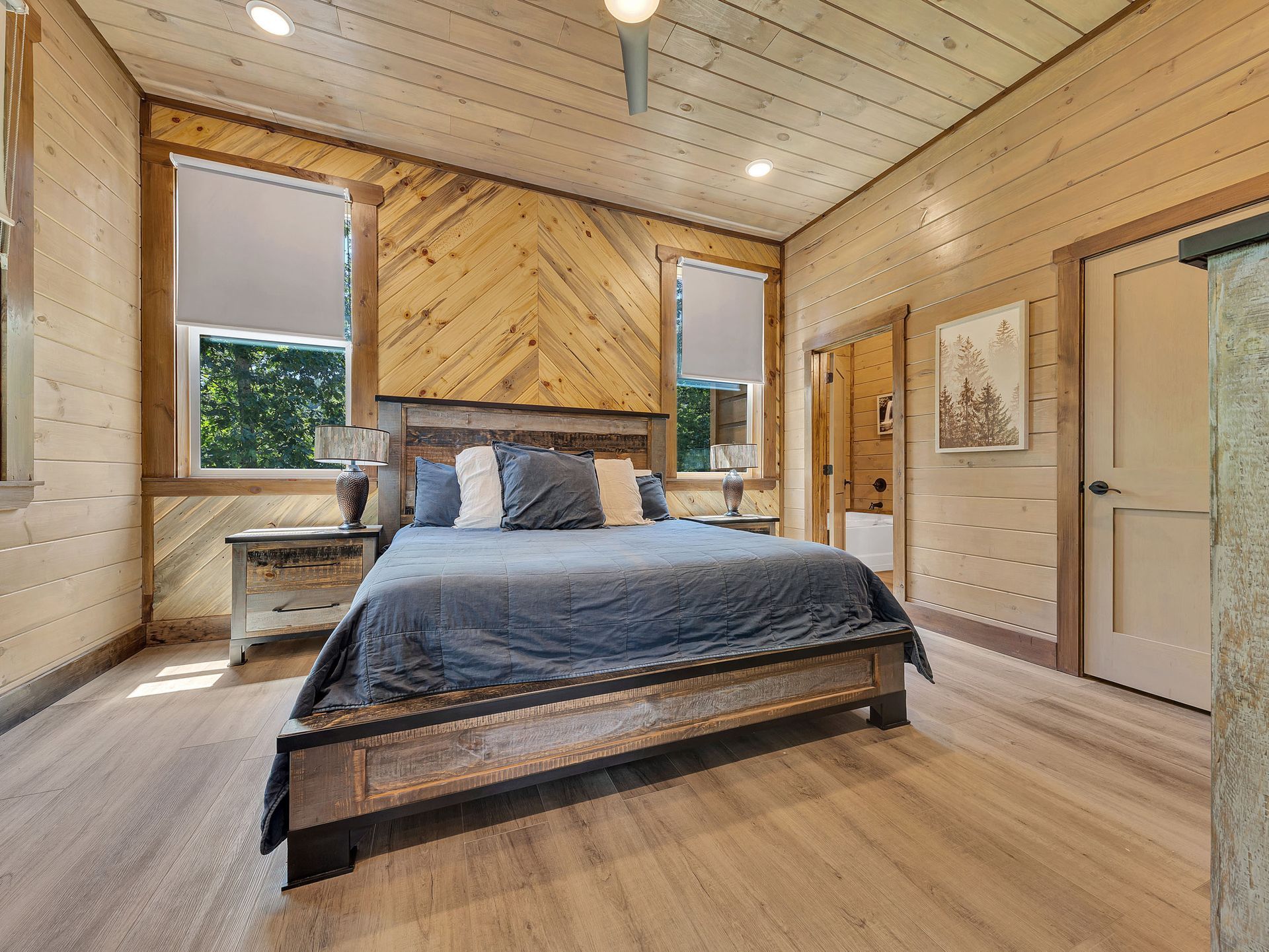 Bedroom with wood paneled walls, a rustic bed, two windows with shades, and a door.