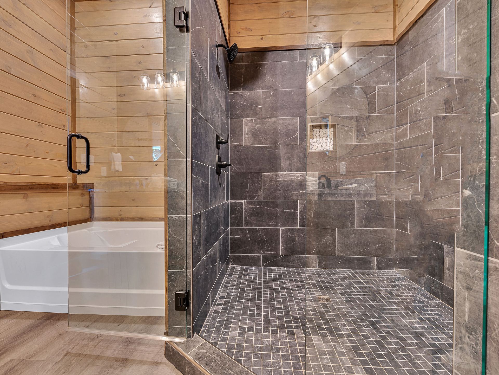 Bathroom with dark gray tiled shower, glass door, white tub, and wood paneling.
