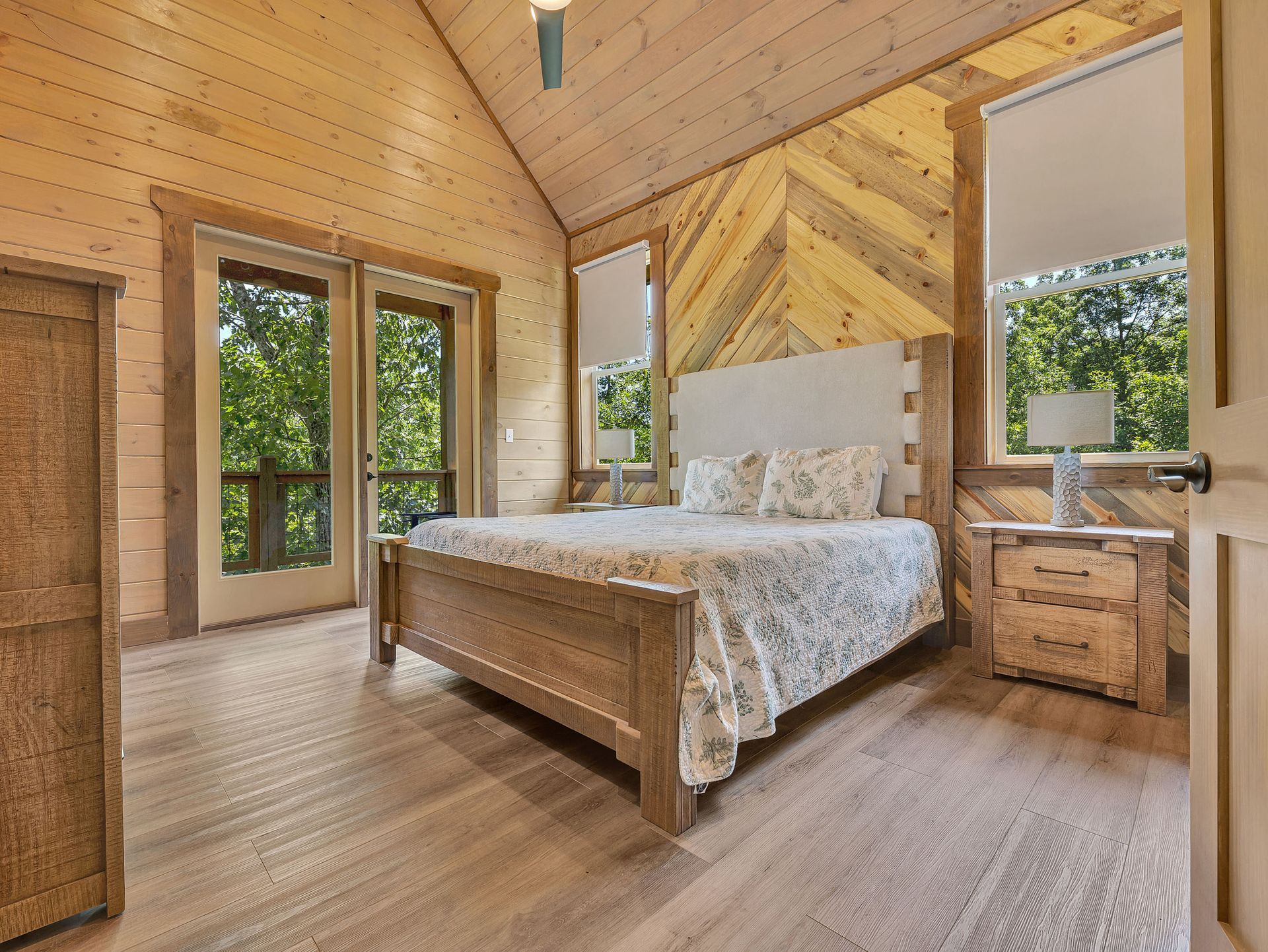Bedroom with light wood walls, bed, and large windows overlooking greenery.