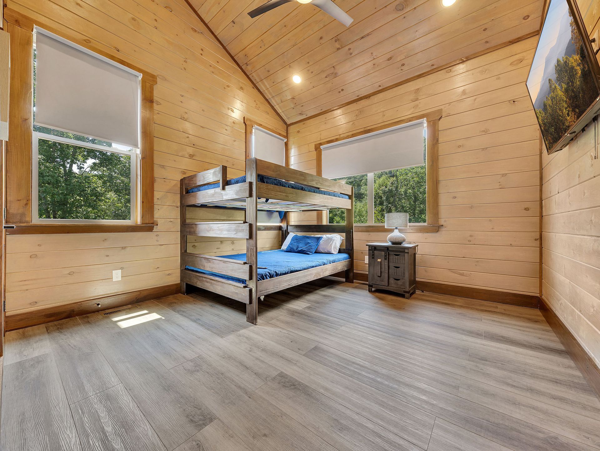 Bunk beds in a wooden-paneled room with windows. Light blue bedding, gray floors, and a TV are present.