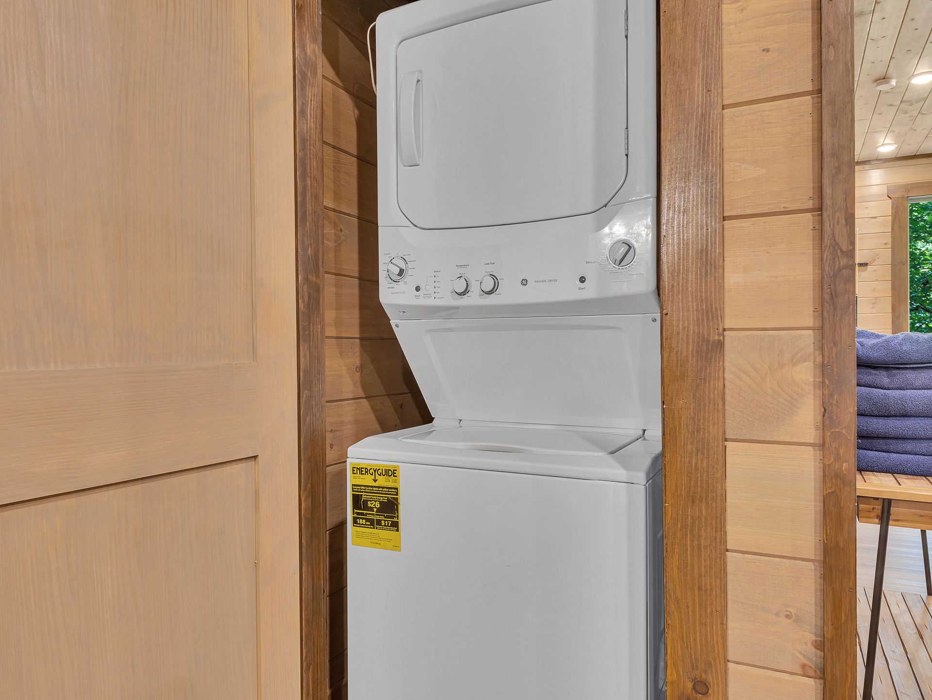 Stacked white washer and dryer unit in a wooden-framed nook, adjacent to a wooden wall and a door.