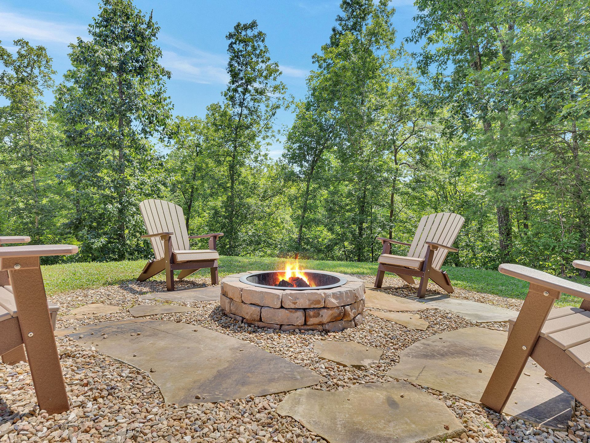 Fire pit with Adirondack chairs surrounded by trees under a blue sky.