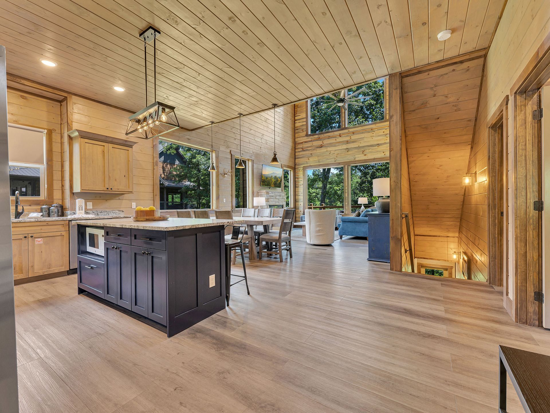 Spacious kitchen with a dark island, light wood cabinetry, and a dining table. Large windows overlook greenery.