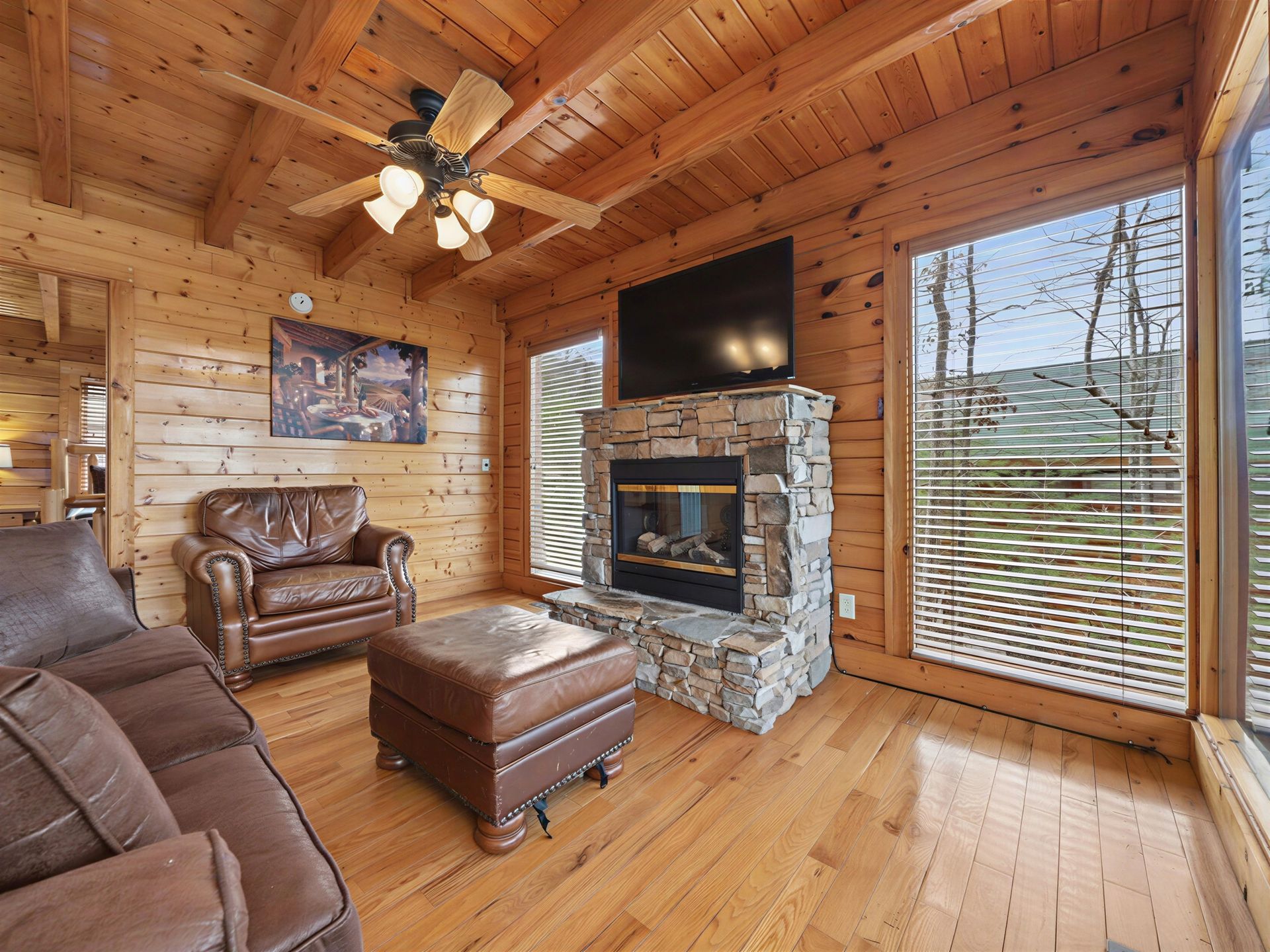 Cozy cabin living room with stone fireplace, TV, leather furniture, and large windows with a view.