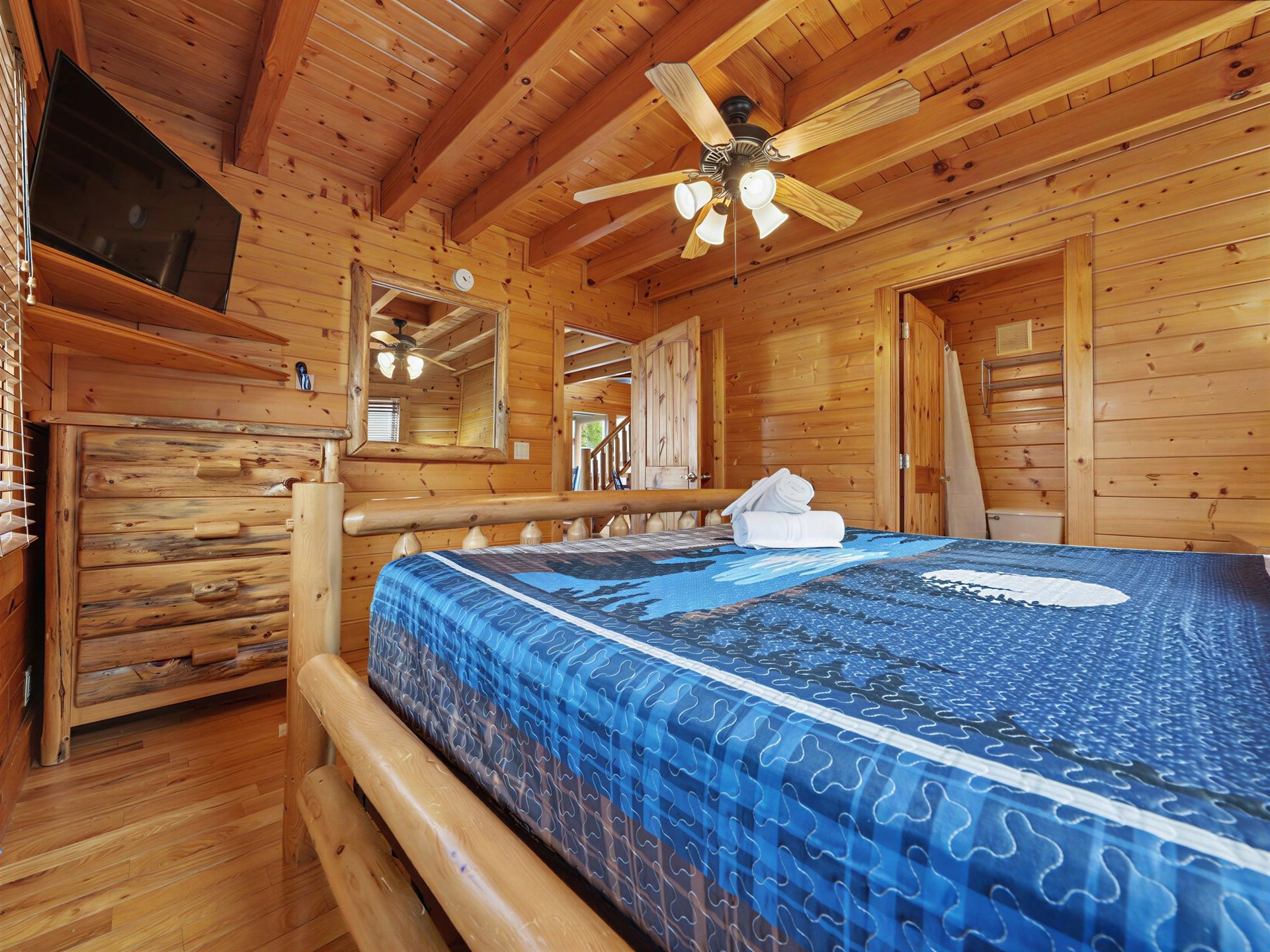 Bedroom in a log cabin with a bed, dresser, TV, and ceiling fan. Wooden walls and flooring.