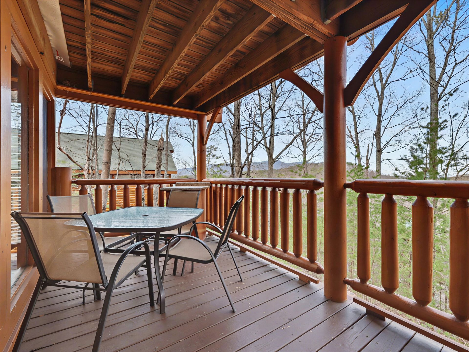 Covered wooden deck with table, chairs, railing, and trees in background.