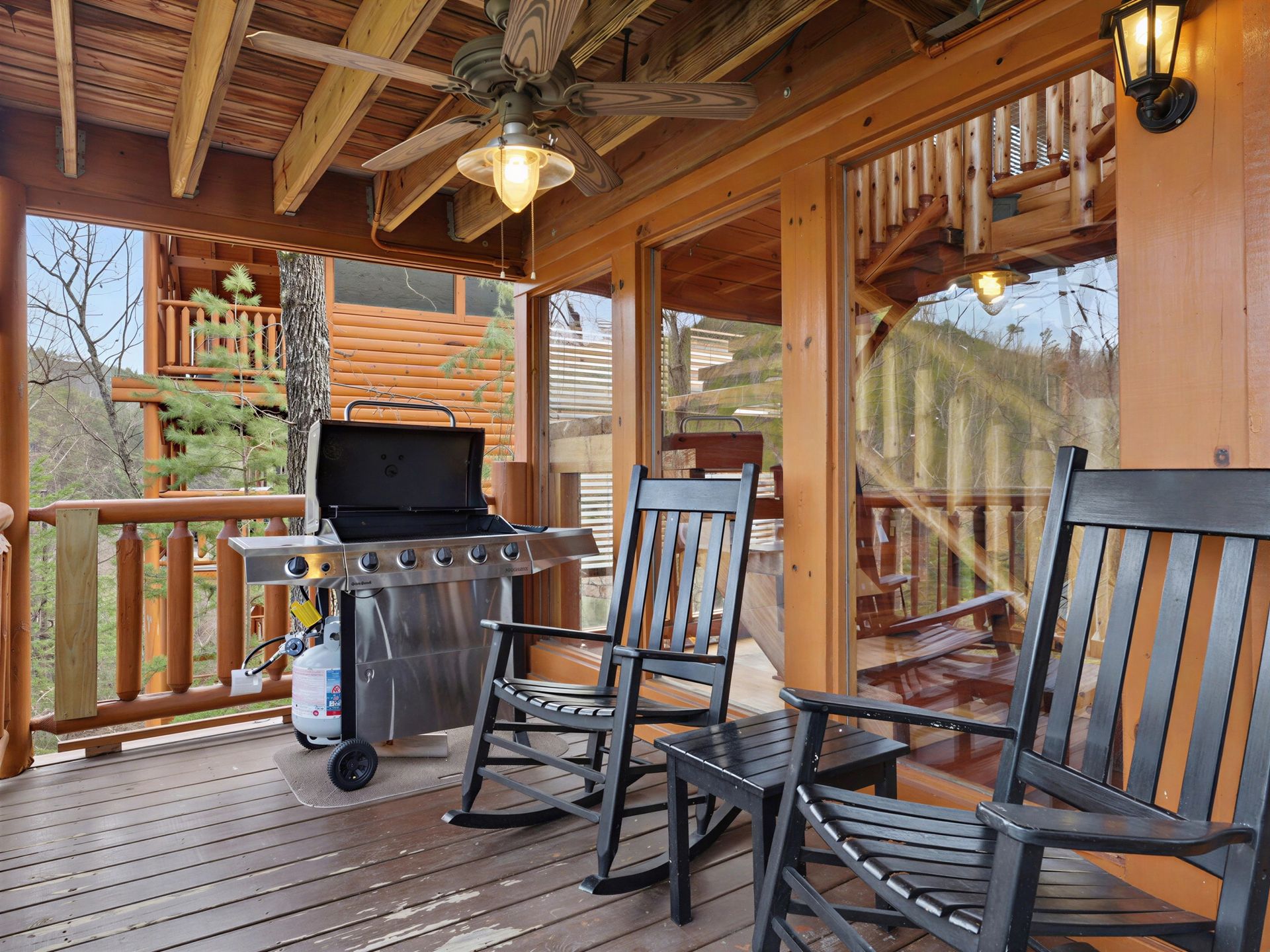 Covered wooden deck with rocking chairs, grill, and view of trees and cabin.