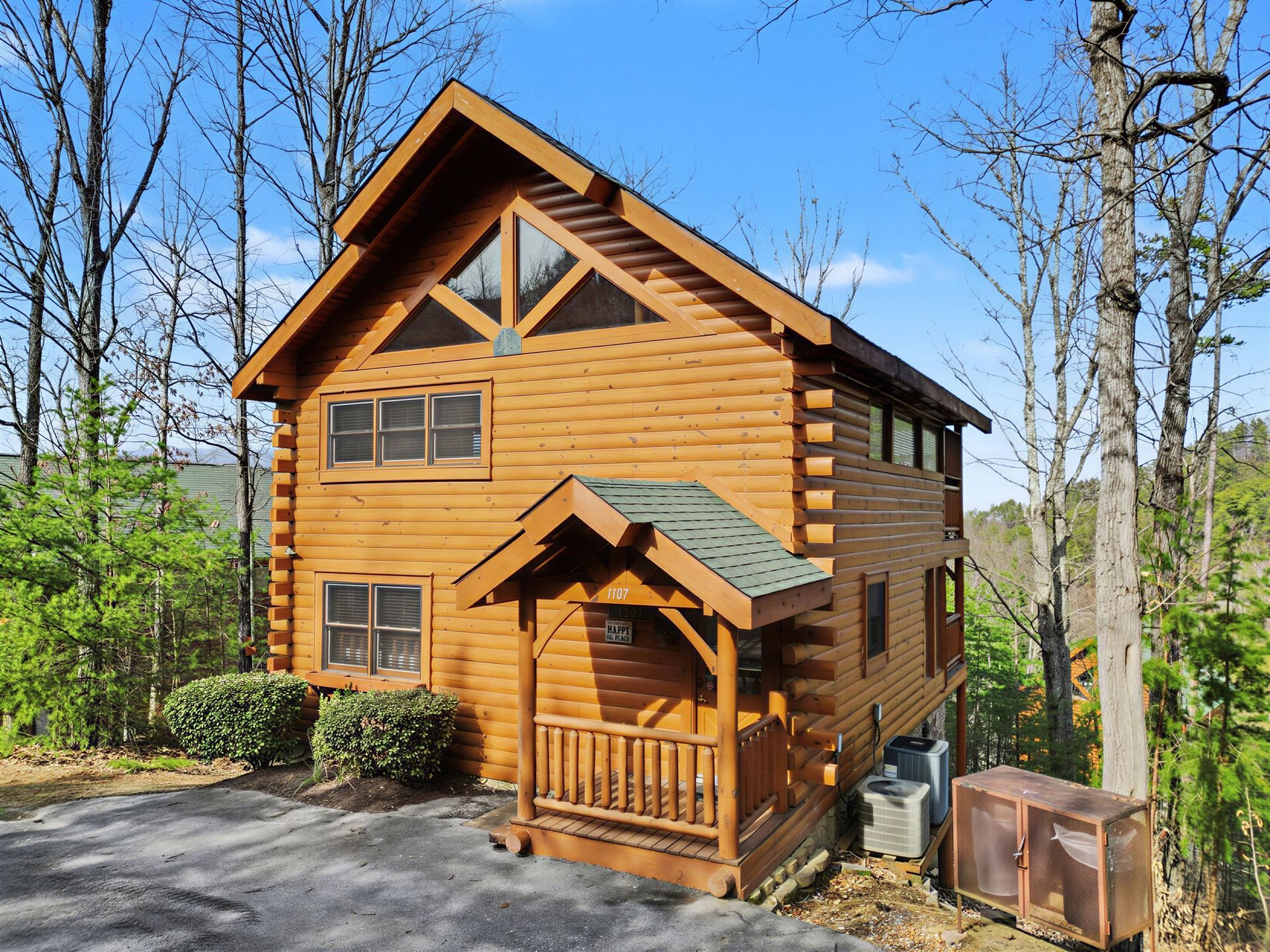 Two-story log cabin with a porch in a wooded setting; brown exterior, green roof, and blue sky.