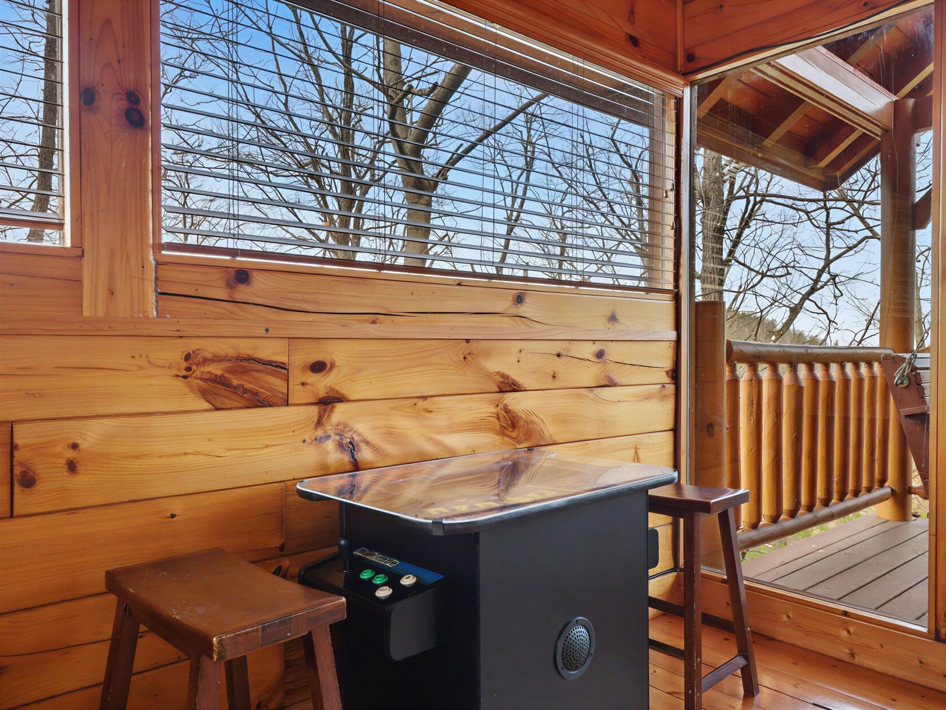 Arcade game table inside a wood-paneled cabin with window views of trees, stool on either side.
